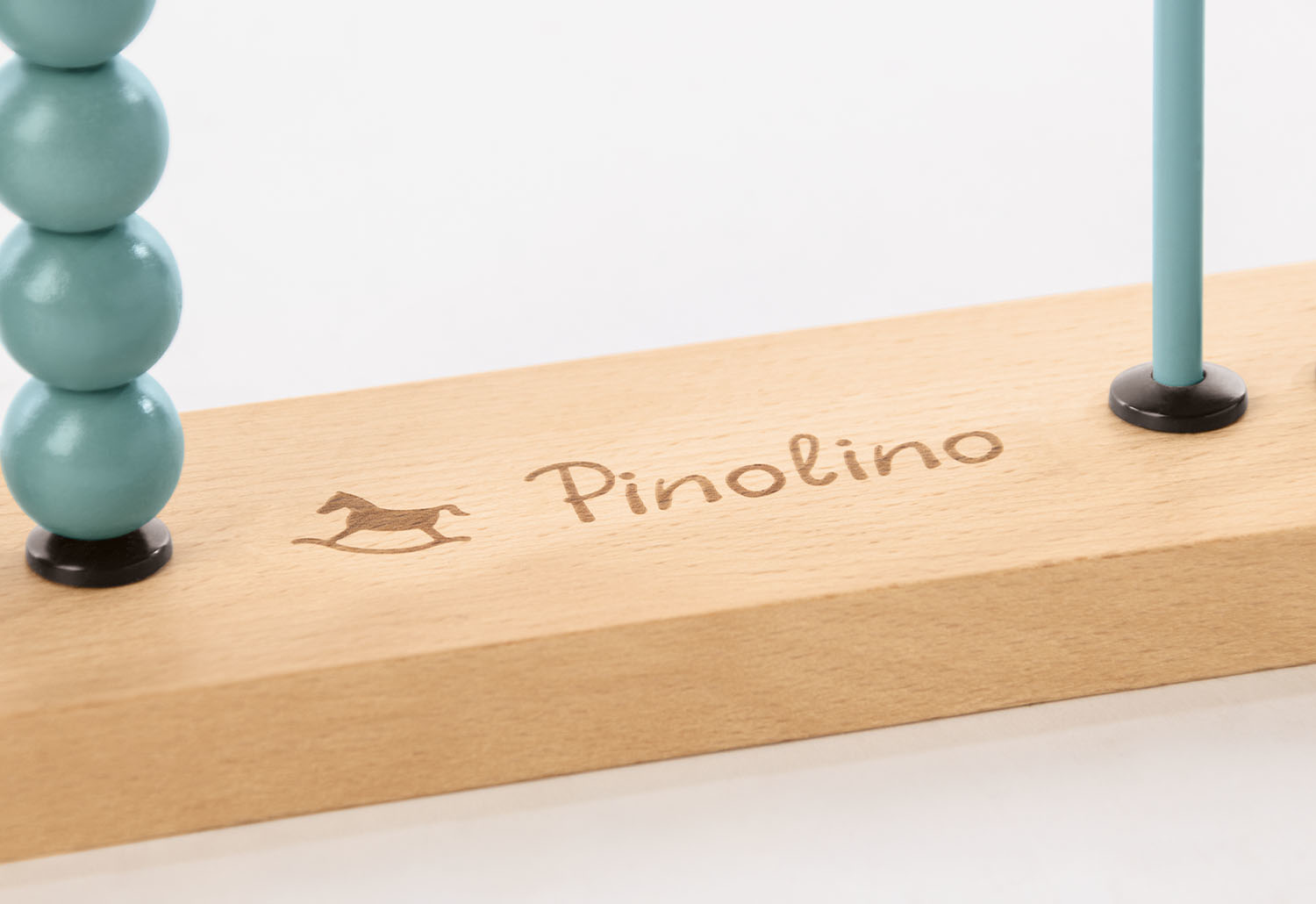 Eine Holzbasis mit der Gravur "Pinolino", einem Schaukelpferd-Symbol und einem blauen Rechenstab mit Perlen.