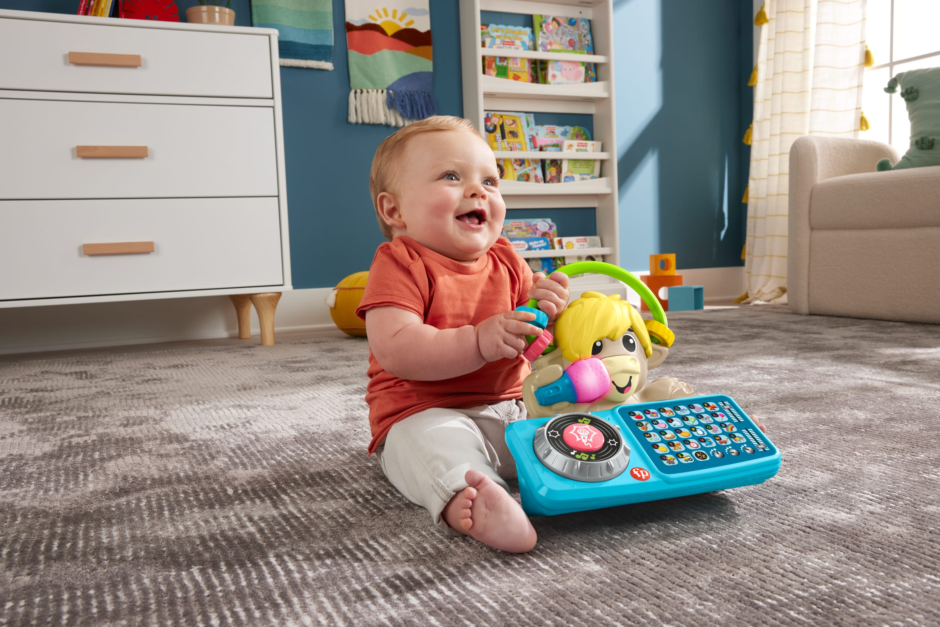 Ein glückliches Baby sitzt auf einem gemusterten Teppich und spielt mit einem blau-gelben DJ-Spielzeug, im Hintergrund eine weiße Kommode und ein Bücherregal.