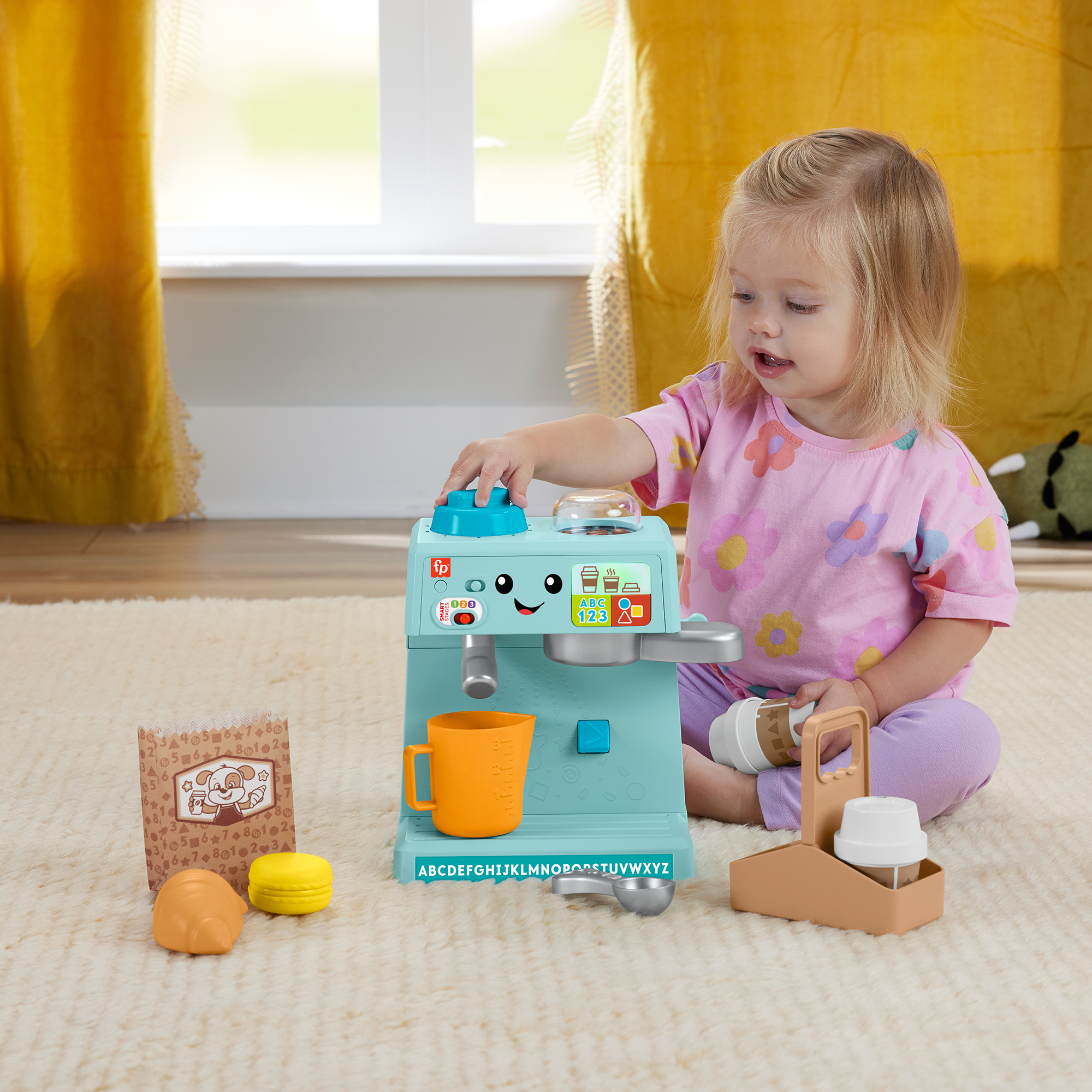Ein kleines Kind in einem pinken Hemd spielt mit einer blauen Spielzeug-Kaffeemaschine, mit Spielzeuggebäck und Tassen in der Nähe.