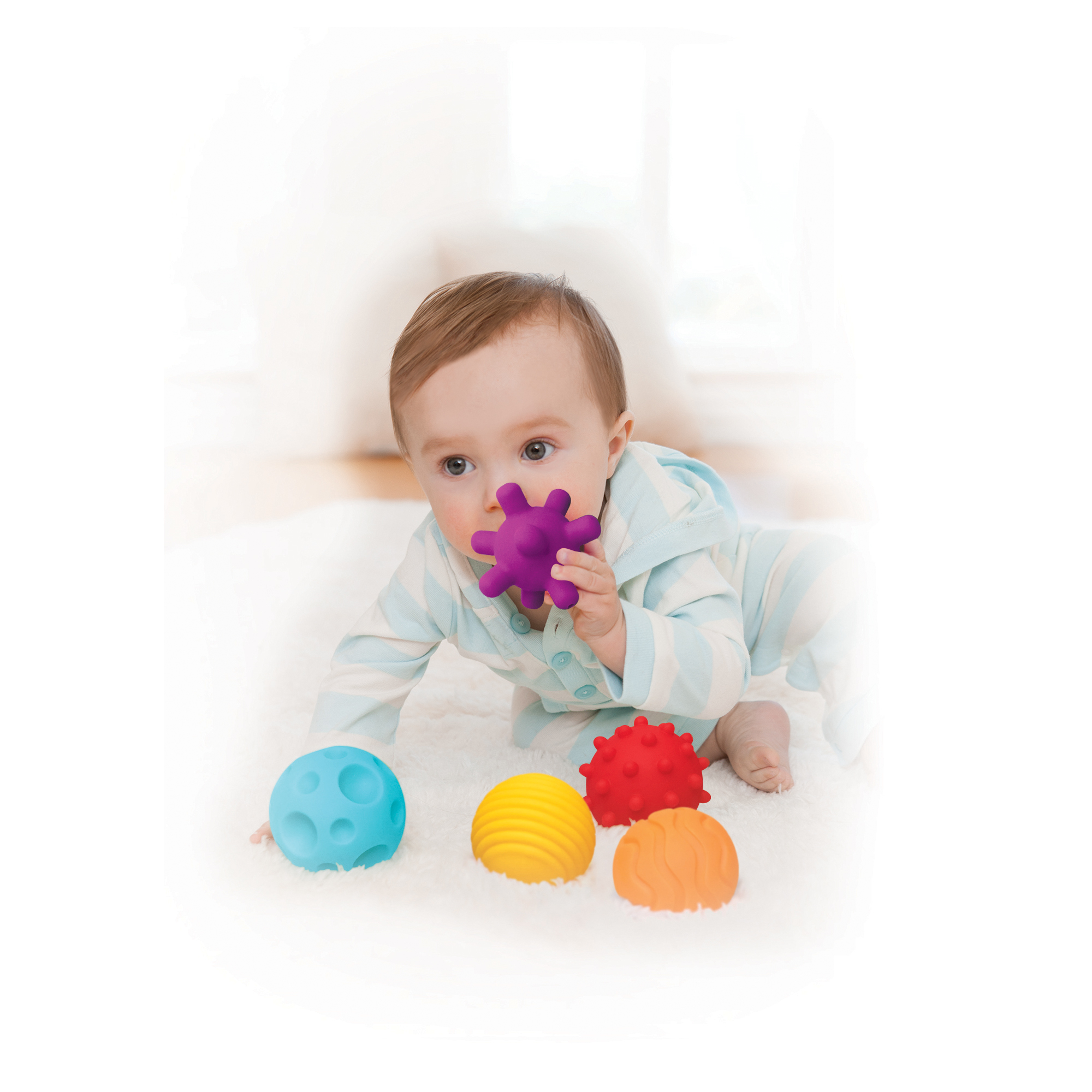 Ein Baby im gestreiften Schlafanzug liegt auf einem weißen Teppich, hält einen lila strukturierten Ball im Mund, umgeben von vier weiteren bunten Bällen.