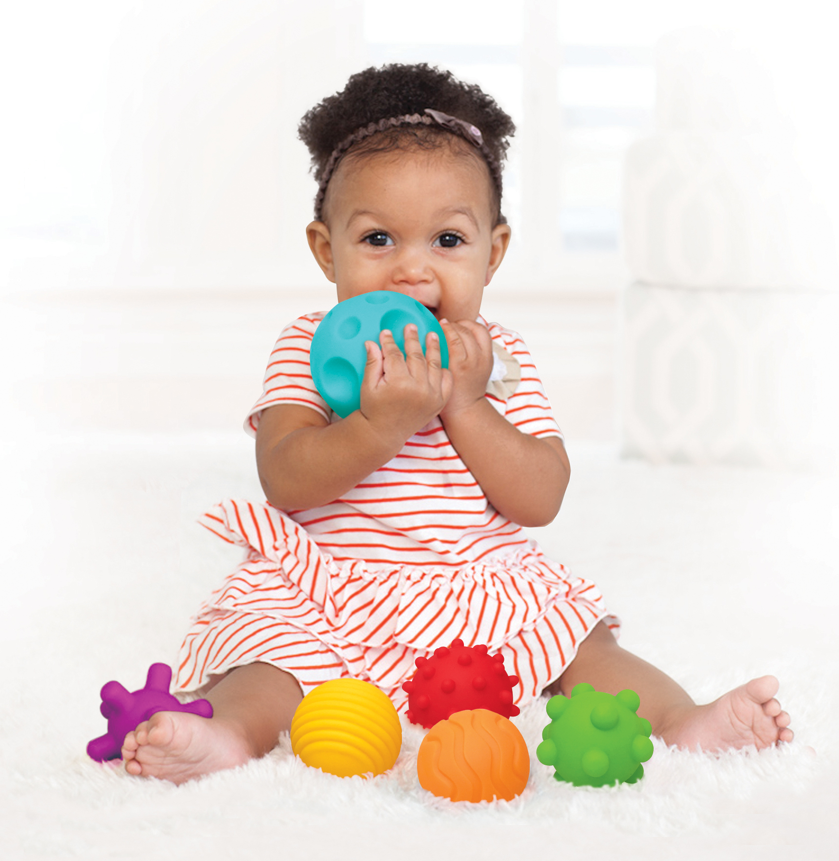 Ein Baby mit einem türkisfarbenen Ball im Mund, umgeben von bunten Spielzeugen auf einem weißen Teppich.