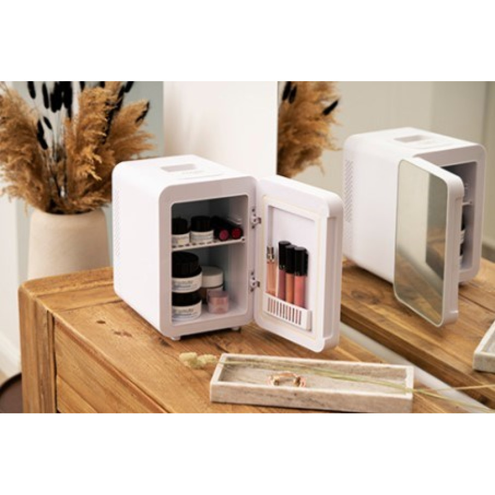 Un mini-frigo bianco su una superficie di legno, la sua porta aperta rivela prodotti per la cura della pelle e lucidalabbra. Un secondo frigo chiuso si riflette in uno specchio dietro.