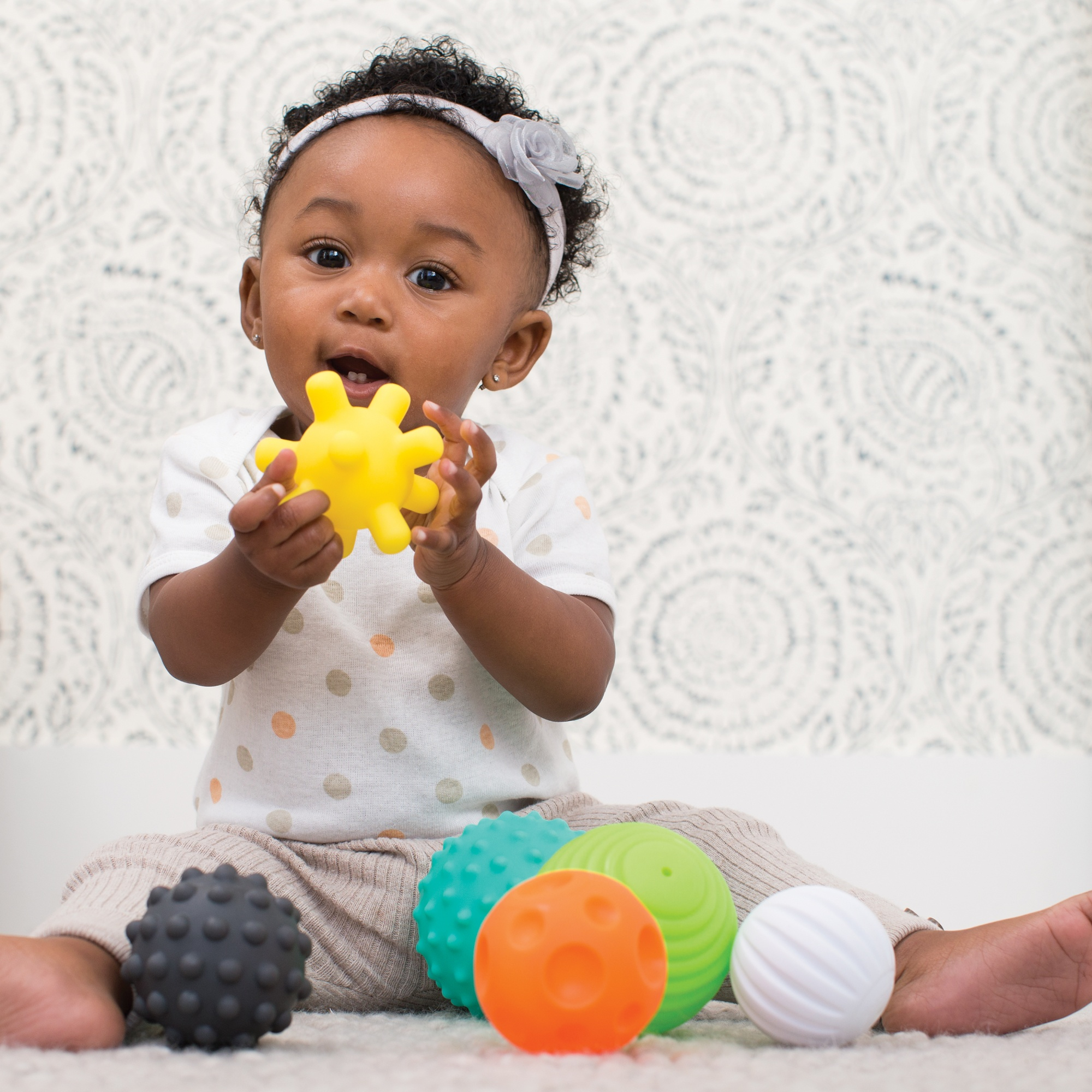 Ein Baby mit einem grauen Stirnband sitzt auf einem Teppich und hält einen gelben Stachelball an den Mund. Andere bunte Bälle liegen verstreut.