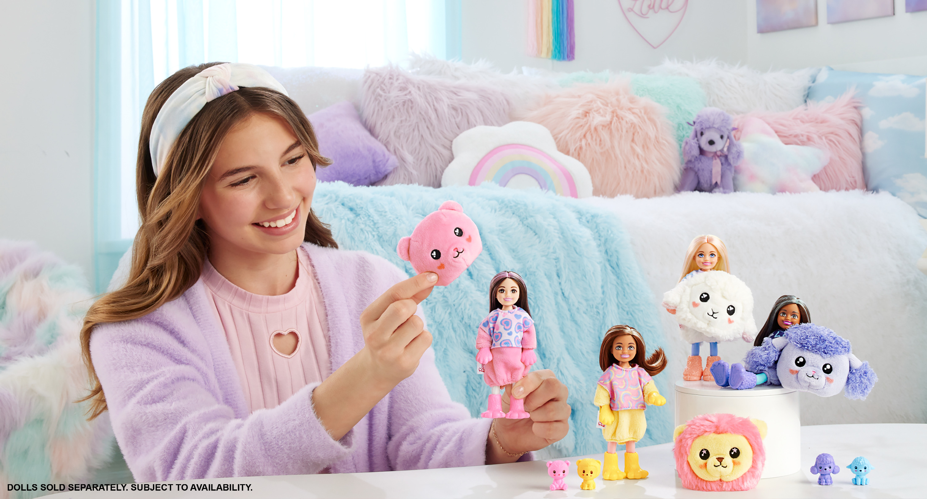Ein lächelndes Mädchen in einem lila Cardigan hält ein pinkes Plüschtier, mit Barbie-Puppen und weiteren Plüschtieren auf einer weißen Oberfläche.