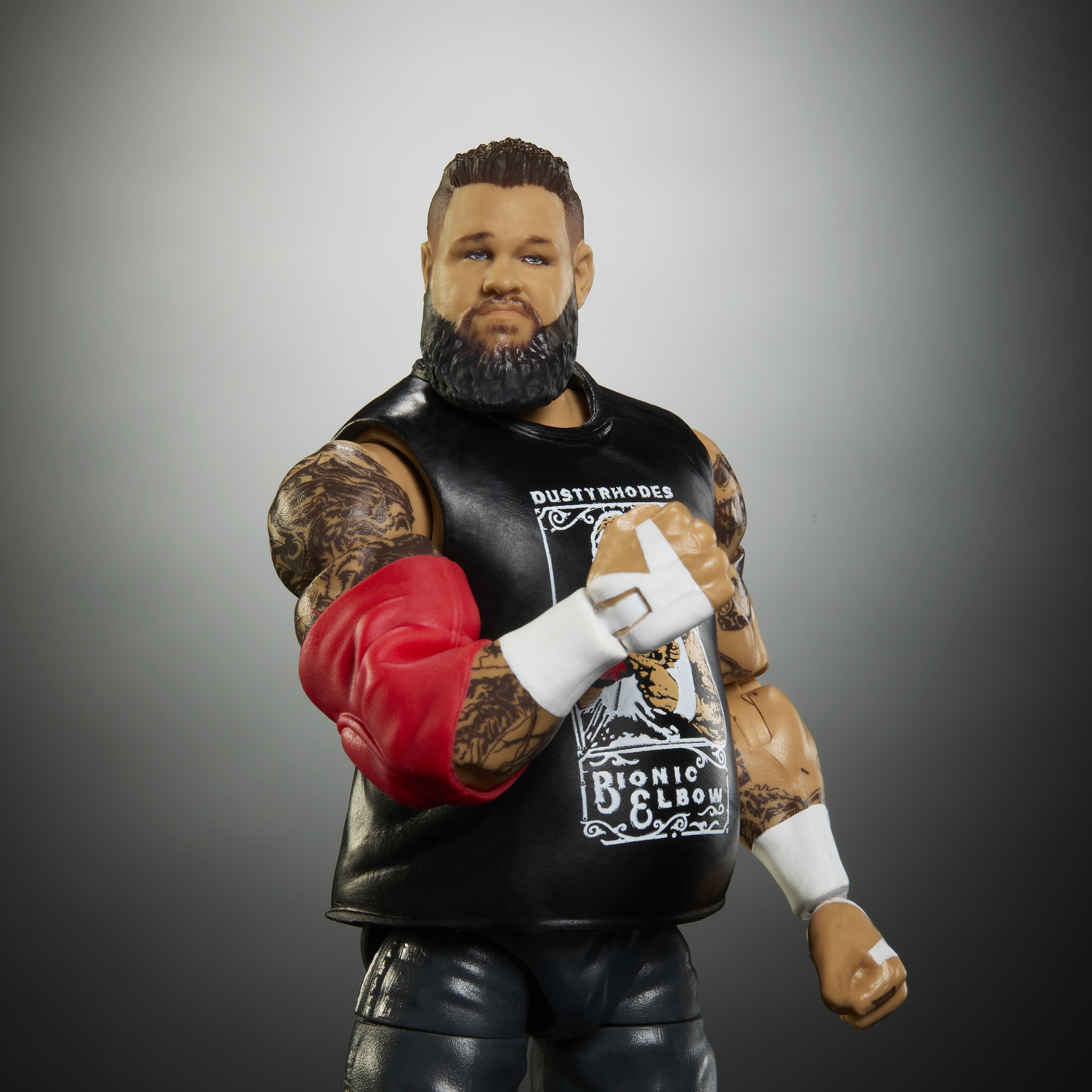 Eine Kevin Owens Actionfigur mit schwarzem Shirt, rotem Ellenbogenschutz und weißem Handgelenksband, stehend vor grauem Hintergrund.