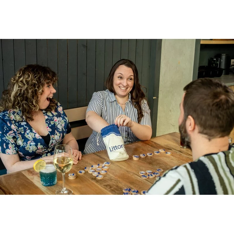Trois personnes à table jouent à un jeu de tuiles. Une femme pioche dans un sac.