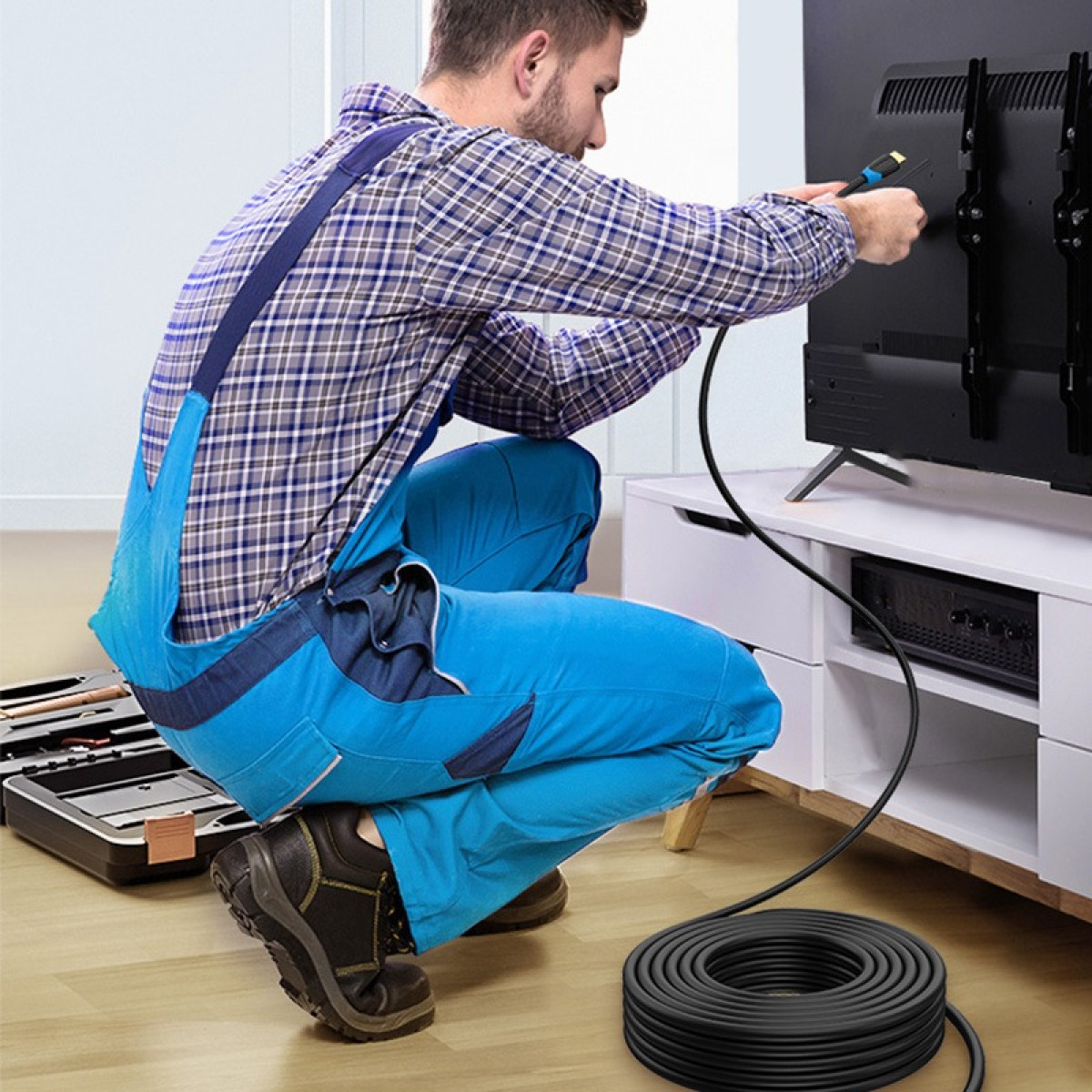 Ein Mann in blauen Overalls und einem karierten Hemd hockt und verbindet ein schwarzes Kabel mit der Rückseite eines schwarzen Fernsehers.