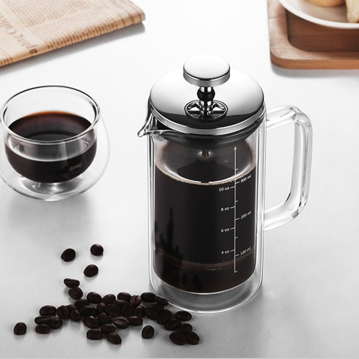 Eine Glas-Kaffeepresse mit Kaffee und eine Tasse daneben, mit Bohnen verstreut.