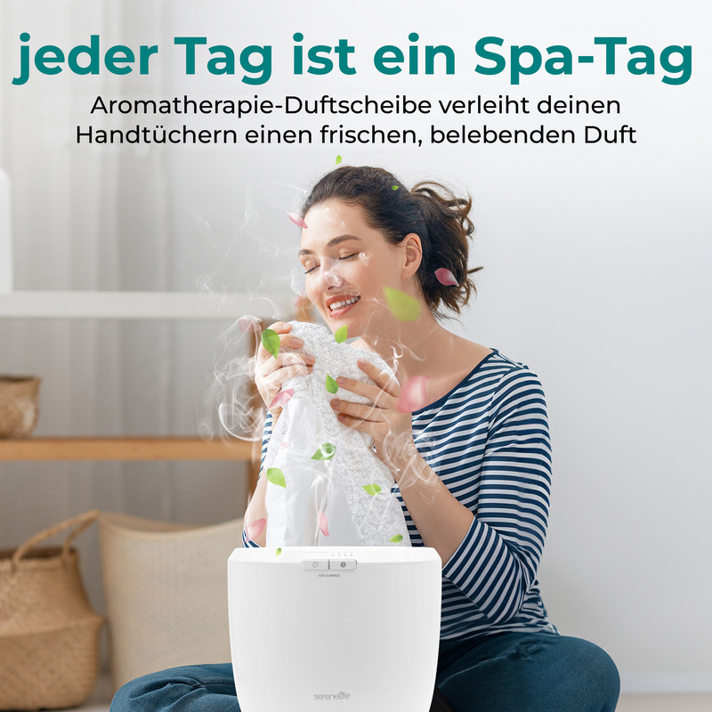 Eine lächelnde Frau in einem gestreiften Hemd atmet den Duft eines weißen Handtuchs ein, während Dampf und Blütenblätter aus einem weißen Gerät aufsteigen.