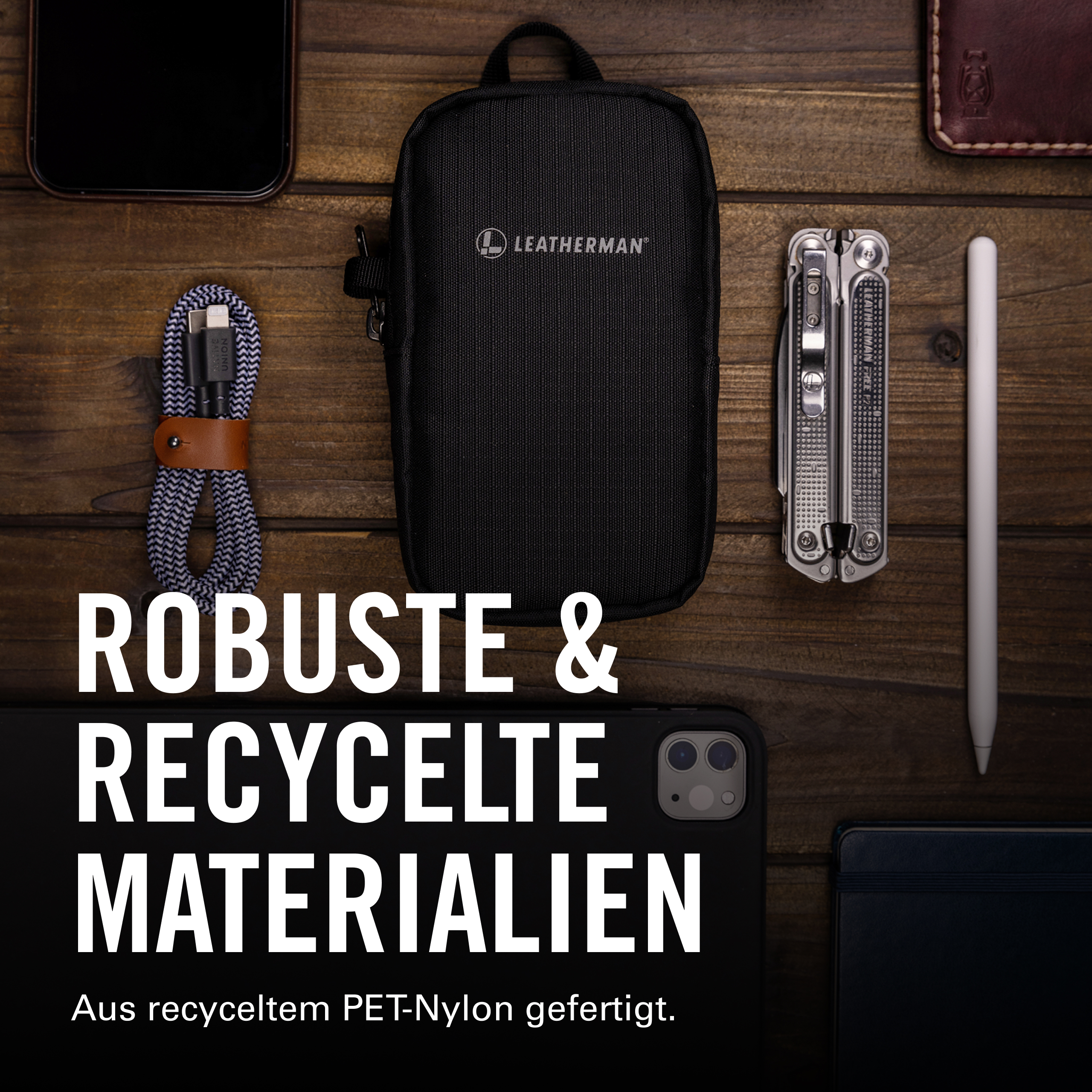 Auf Holzoberfläche liegen ein Handy, Ladekabel, Leatherman-Tasche, Multitool, Stift & Notizbuch.
