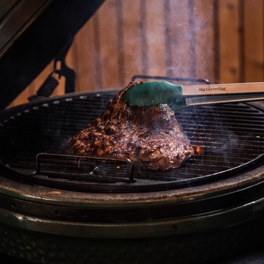 Bir biftek, maşa ile tutularak Big Green Egg'de ızgara yapılıyor. Duman yükseliyor.
