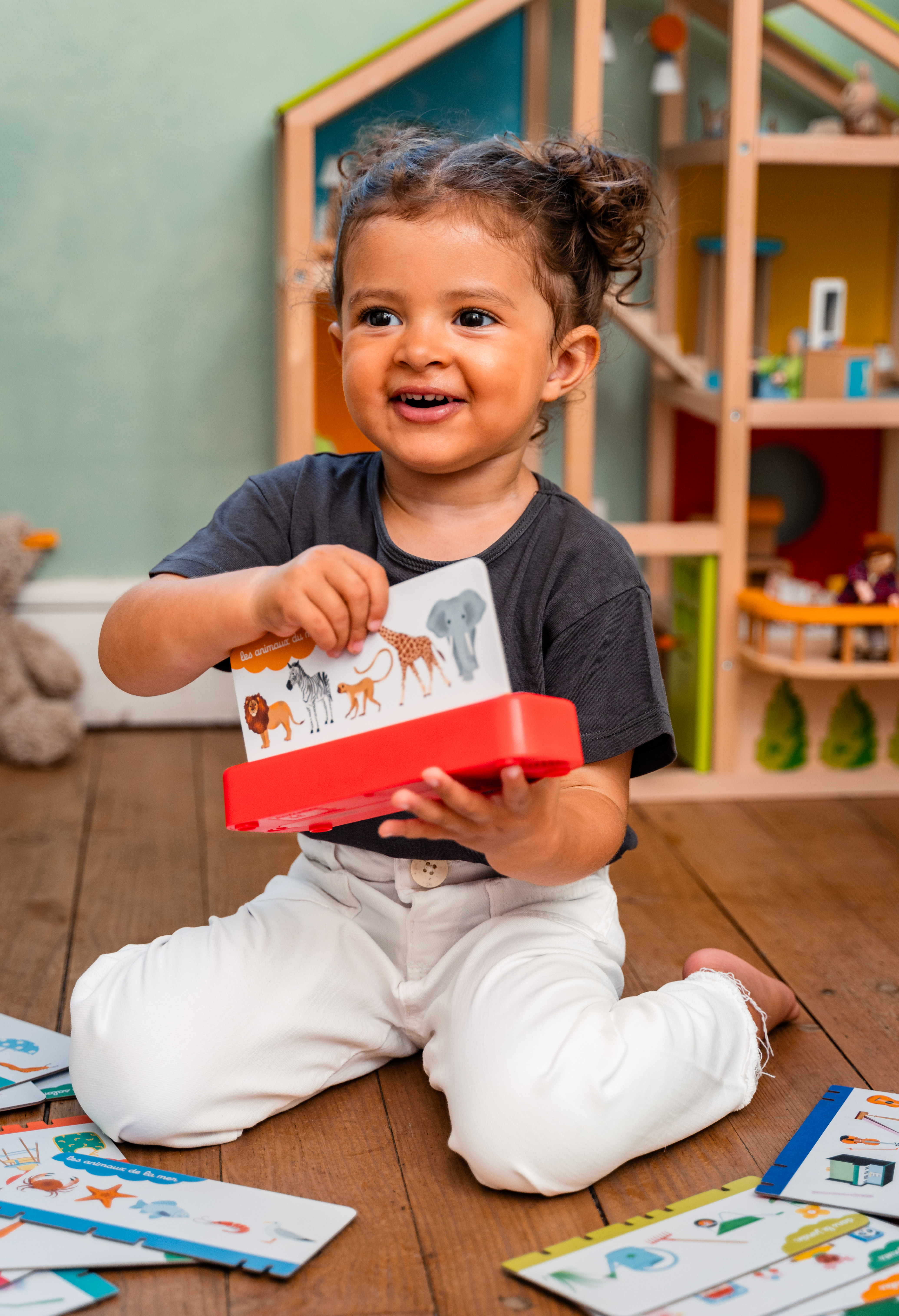 Ein lächelndes Kind mit dunklen Locken sitzt auf einem Holzboden und hält ein rotes Spielzeug mit Tierbildern.
