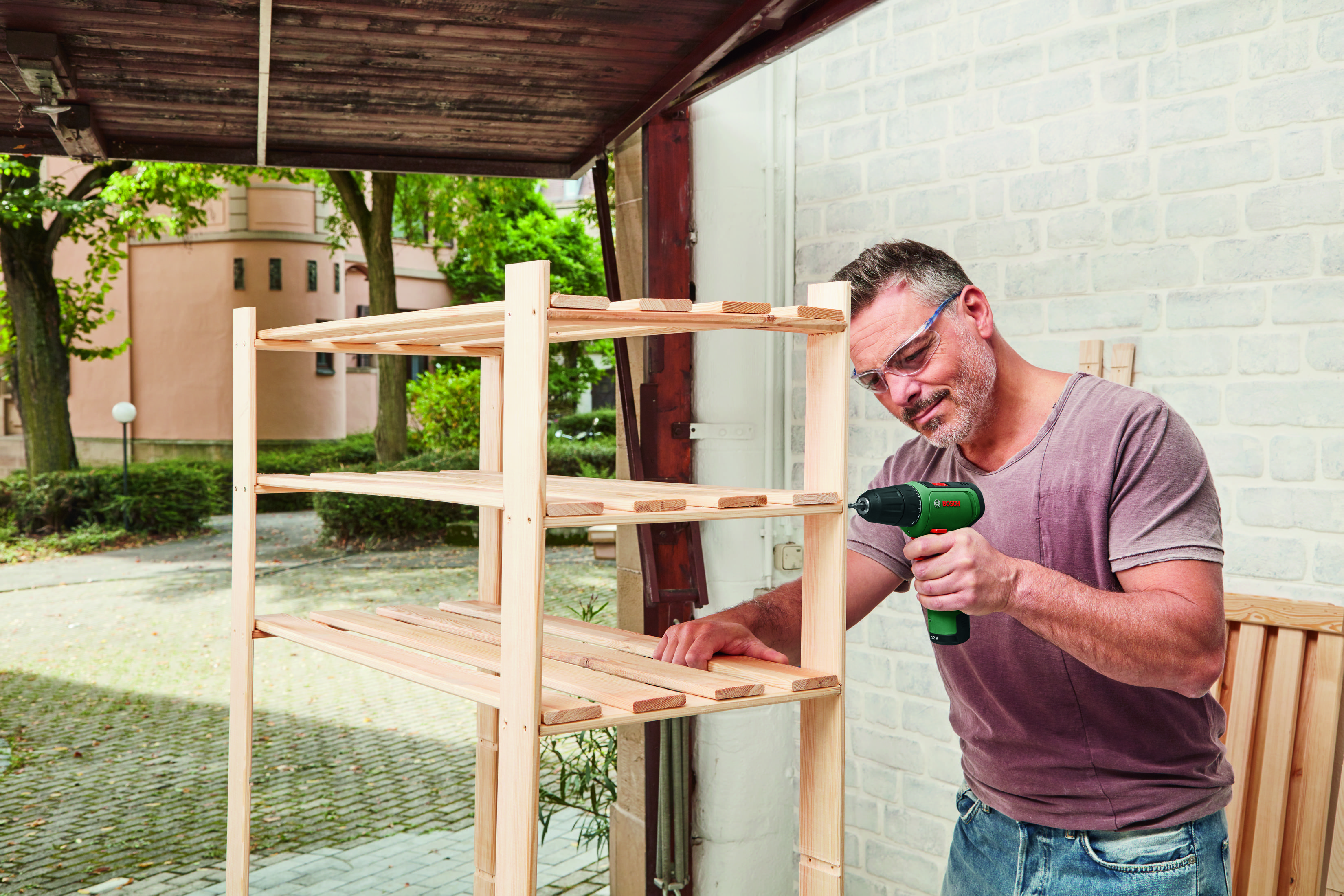 Ein Mann mit Schutzbrille bohrt mit einem Bohrer in ein Holzregal in einer Werkstatt mit einer Ziegelwand.