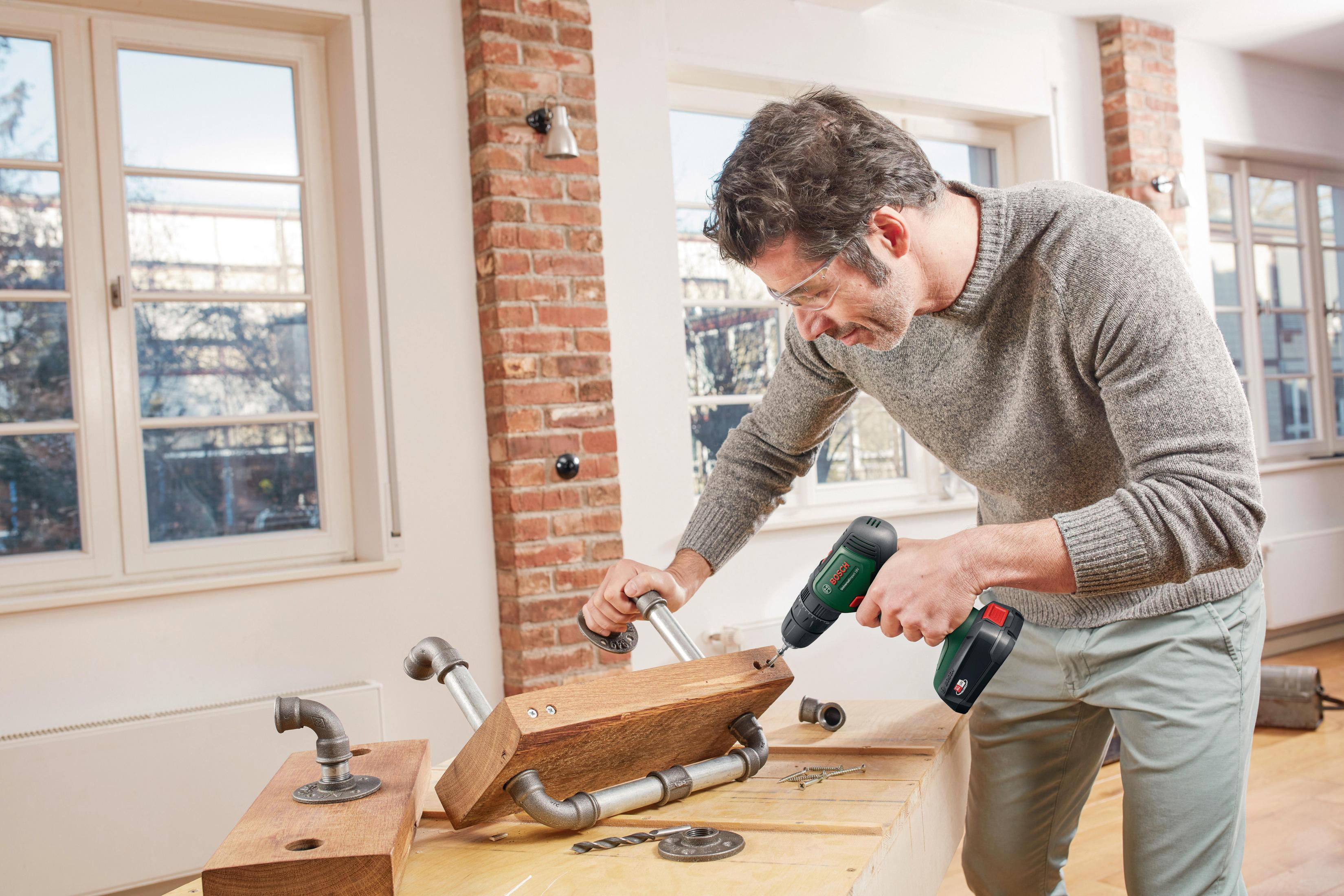Ein Mann mit Schutzbrille benutzt einen Bosch-Bohrer an einem Holzbrett mit Rohrverbindungen, um ein Regal zusammenzubauen.