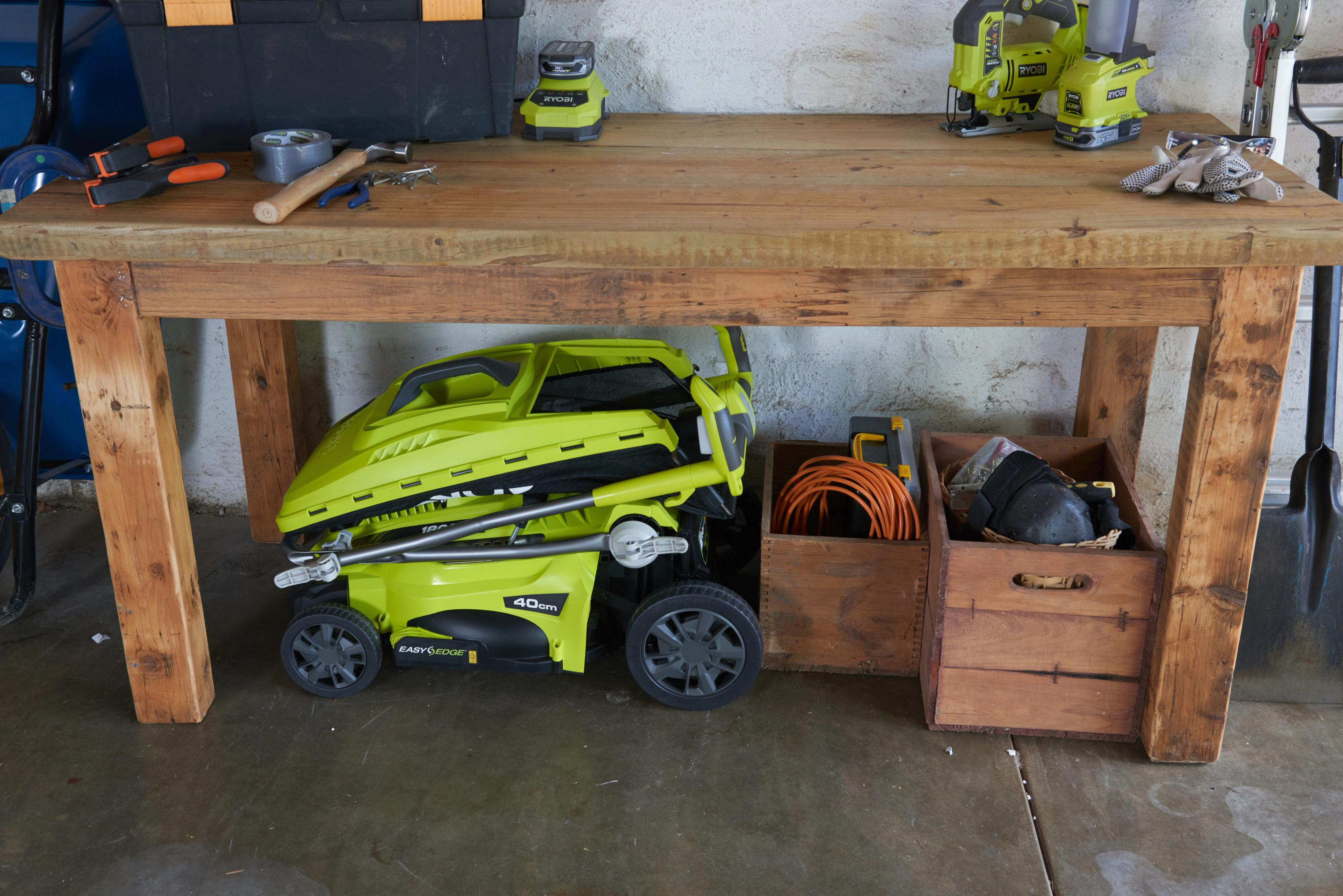 Eine hölzerne Werkbank in einer Garage mit Werkzeugen oben und einem zusammengeklappten lindgrünen Rasenmäher darunter, neben Holzkisten.