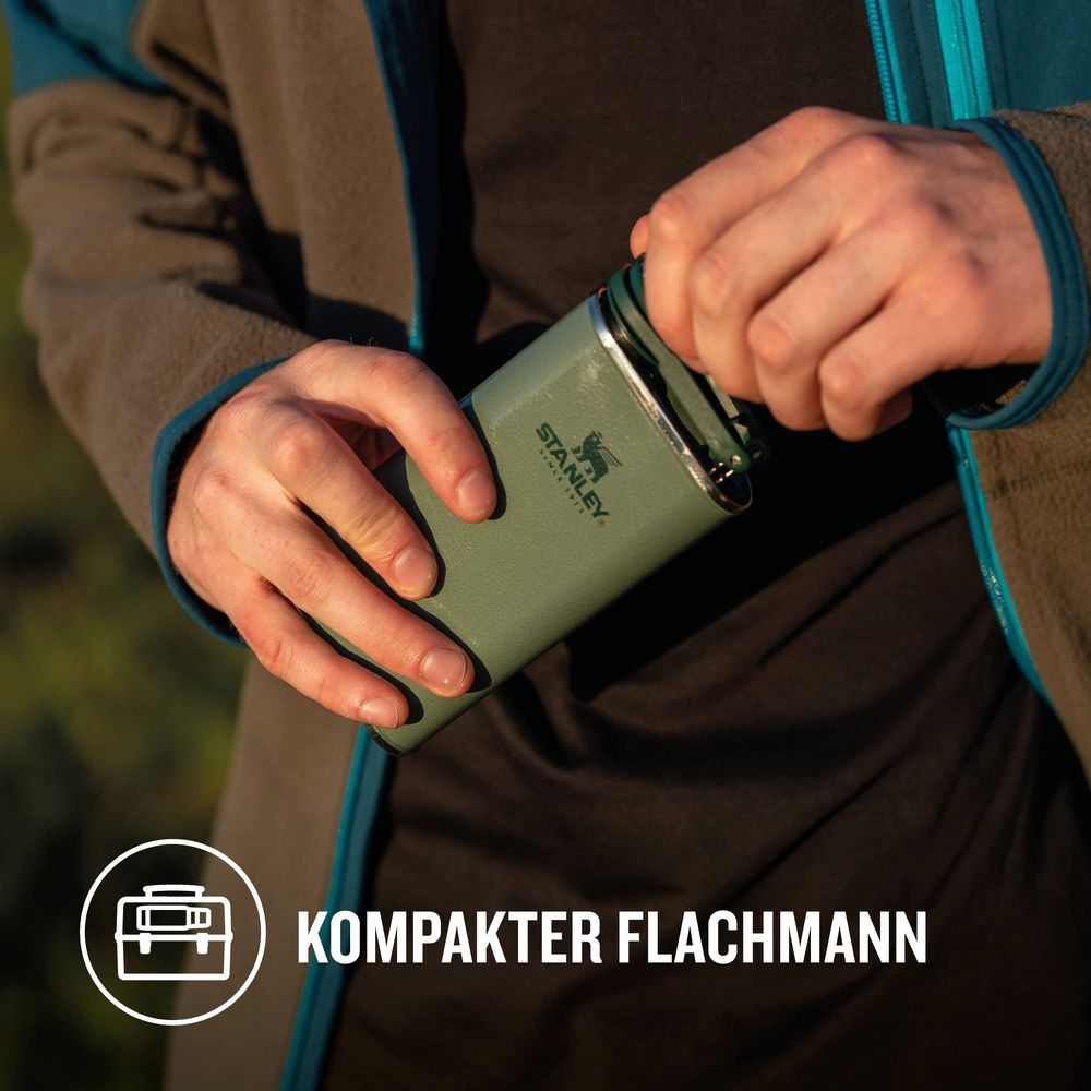 Eine Person in einer braun-blauen Jacke hält einen grünen Stanley-Flachmann und öffnet dessen Verschluss. "KOMPAKTER FLACHMANN" ist sichtbar.