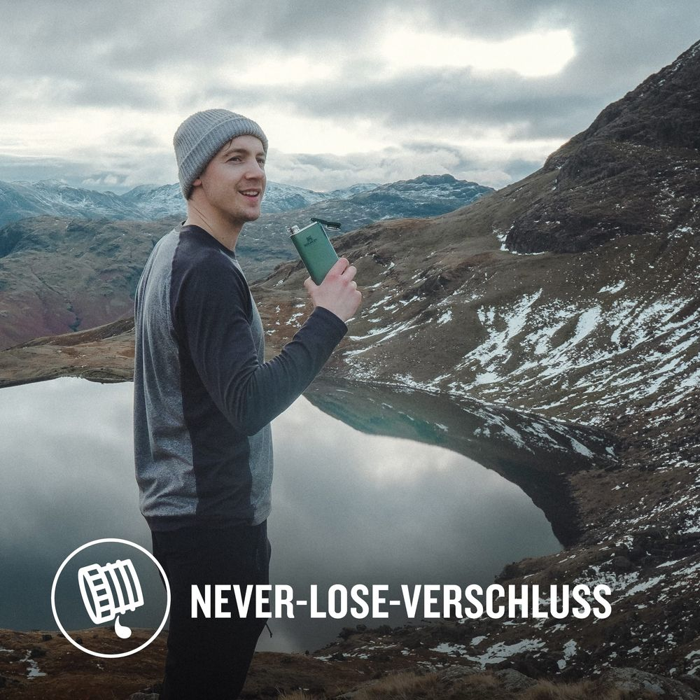 Ein Mann mit grauer Mütze und Hemd hält eine grüne Flasche in einer verschneiten Berglandschaft mit See.