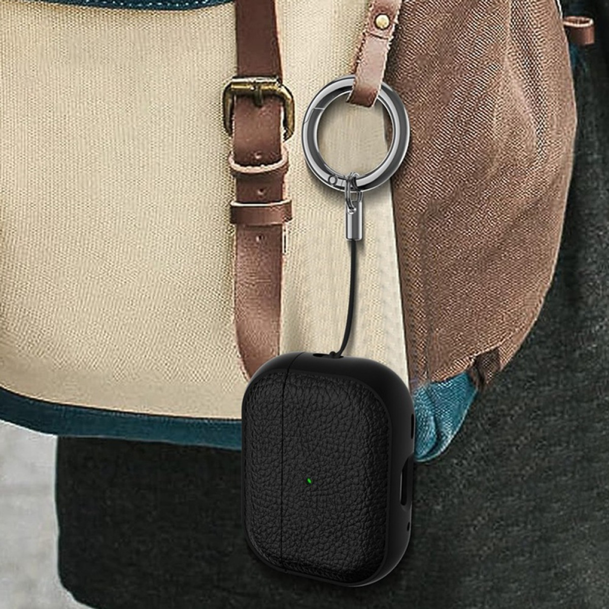 Ein schwarzes strukturiertes AirPods-Etui mit grünem Licht, an einem silbernen Schlüsselanhänger befestigt, hängt an einem beige-braunen Rucksack.