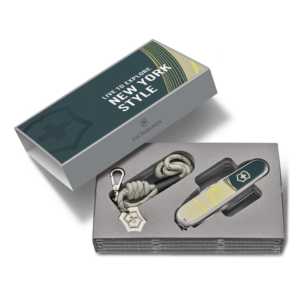 Beyaz arka planda açık bir Victorinox hediye kutusu, içinde çok amaçlı bir alet ve gri ip ve metal klipsli bir anahtarlık bulunuyor.