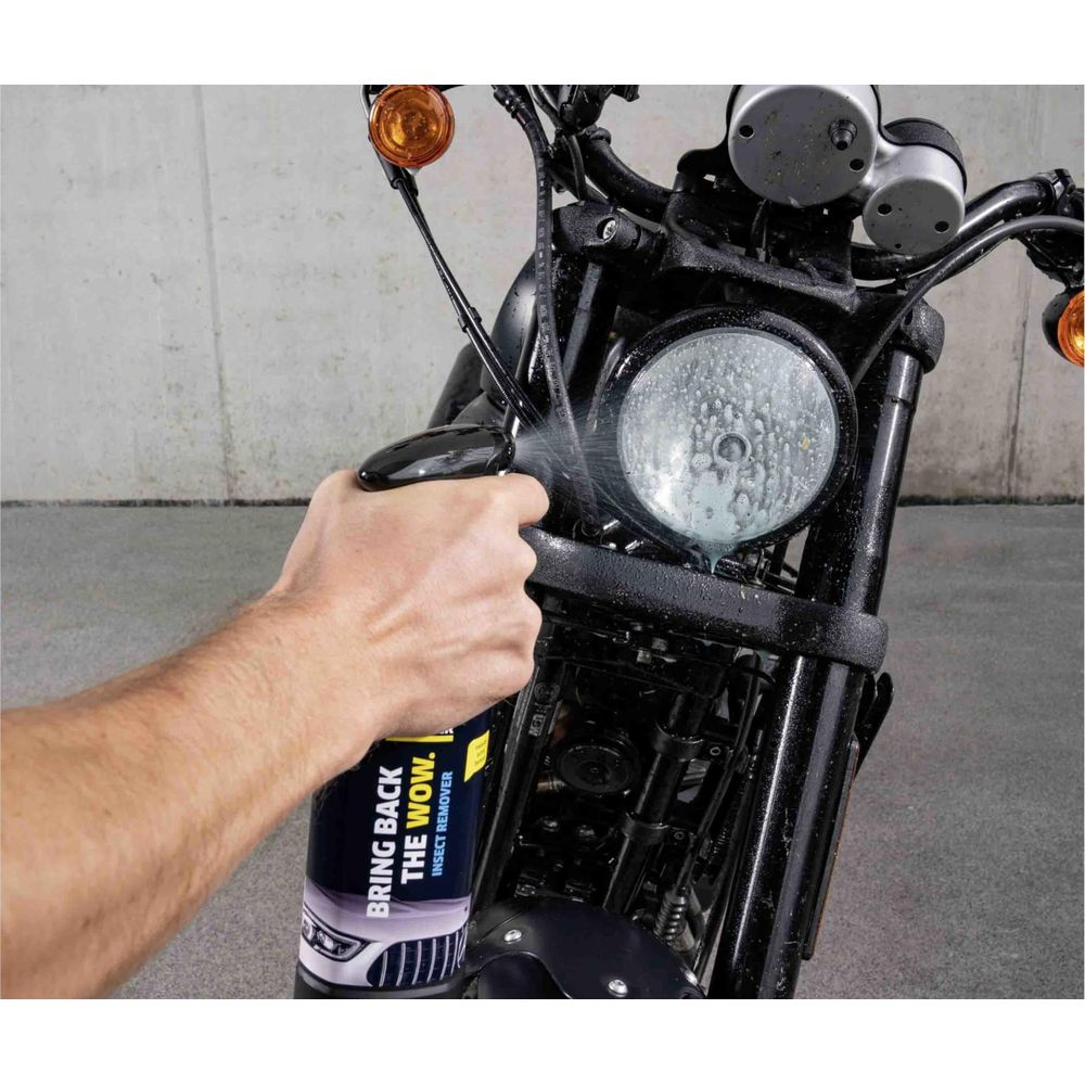 Eine Person sprüht einen schwarzen Motorradscheinwerfer mit einer Reinigungsflasche mit der Aufschrift „BRING BACK THE WOW. INSECT REMOVER.“ ein.