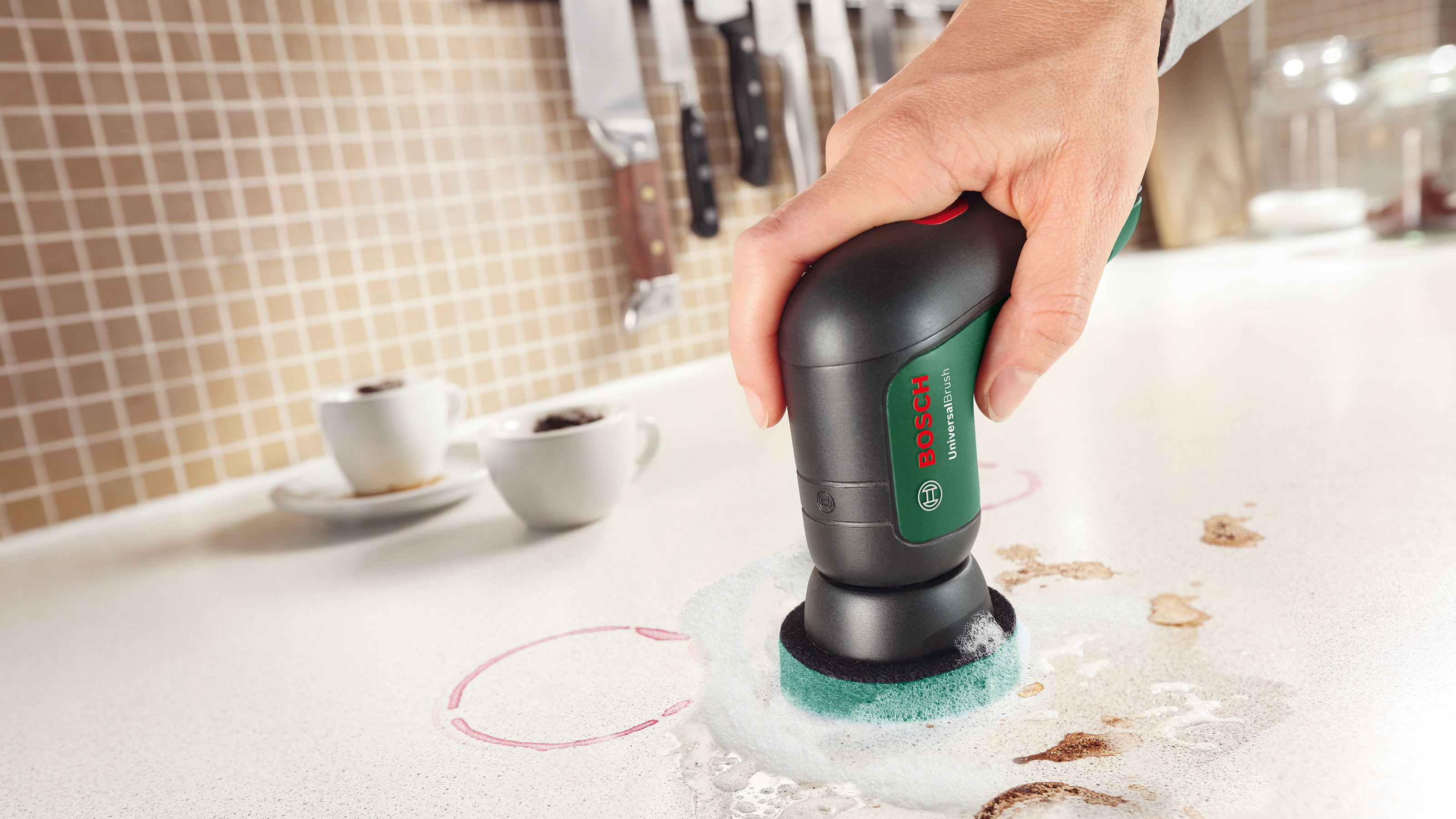 Eine Hand hält eine Bosch UniversalBrush und reinigt eine Küchenarbeitsplatte mit einem grünen Scheuerschwamm und weißem Schaum.