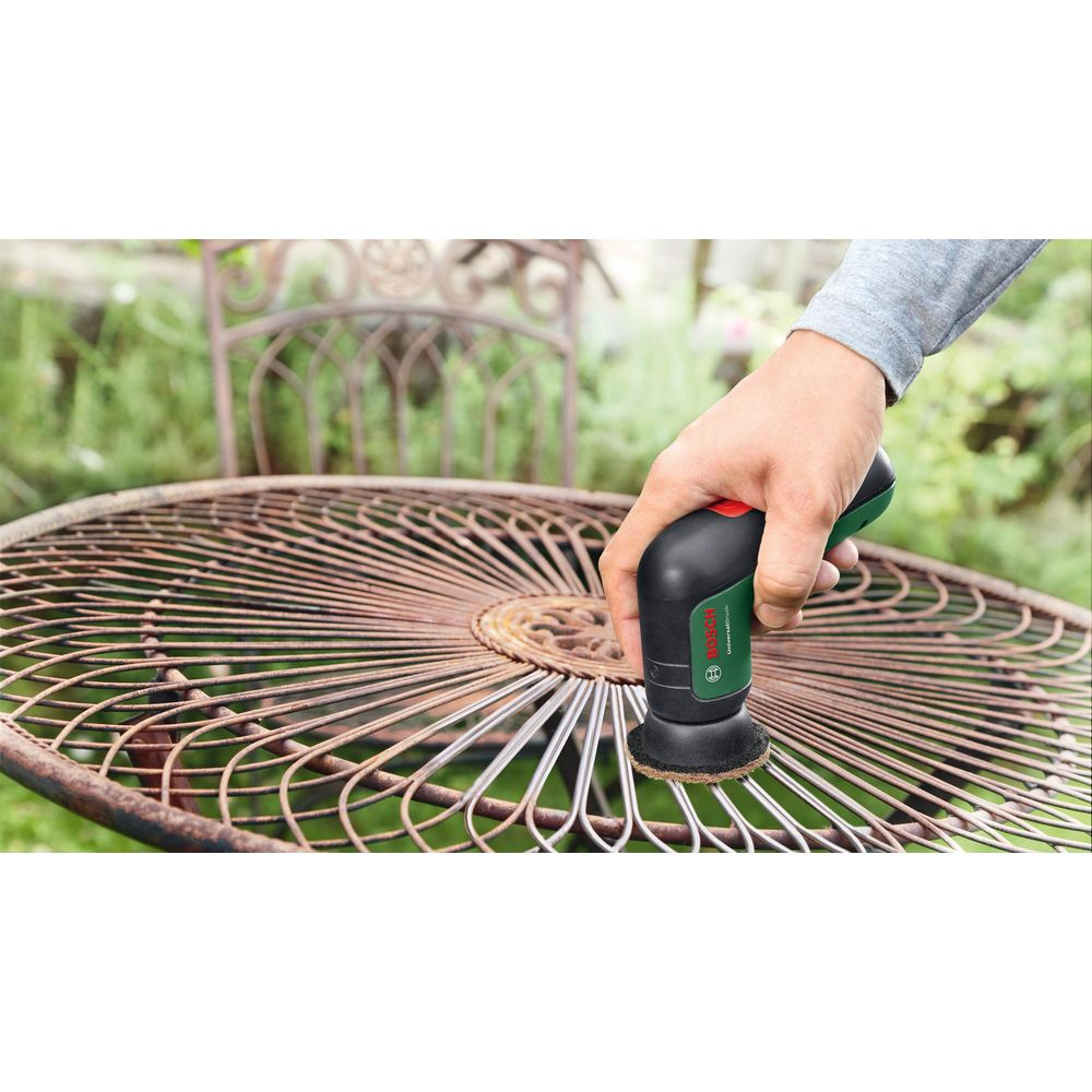 Eine Person benutzt eine Bosch UniversalBrush, um einen rostigen Metallgartentisch zu reinigen, mit einem weiteren rostigen Stuhl im Hintergrund.