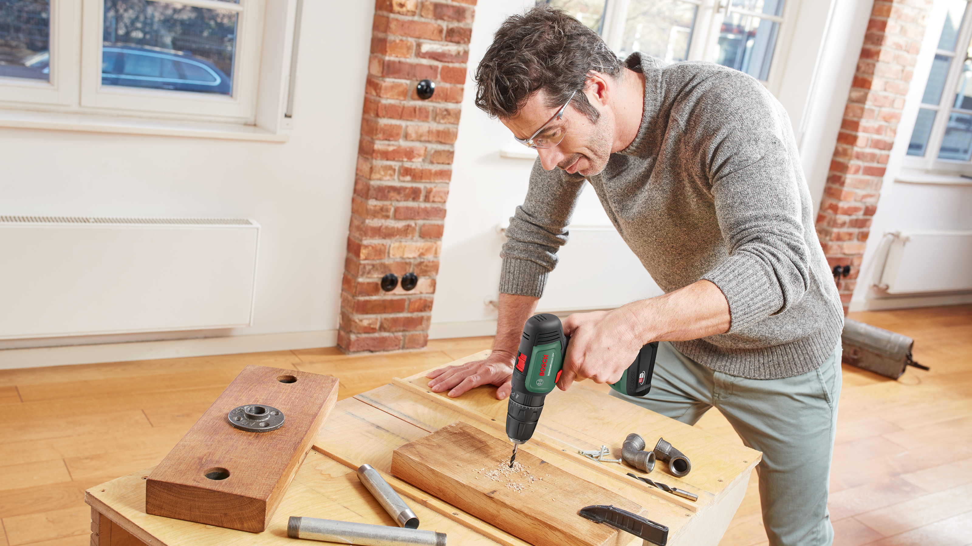 Ein Mann mit Schutzbrille bohrt in ein Holzstück auf einer Werkbank, umgeben von Werkzeugen und Holzspänen.