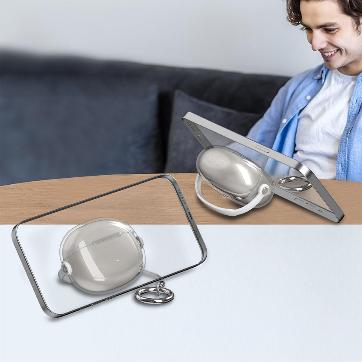 Ein lächelnder Mann schaut auf ein Telefon, das von einem weißen Ohrhörer-Etui auf einem Tisch gestützt wird; ein weiteres Etui im Vordergrund.
