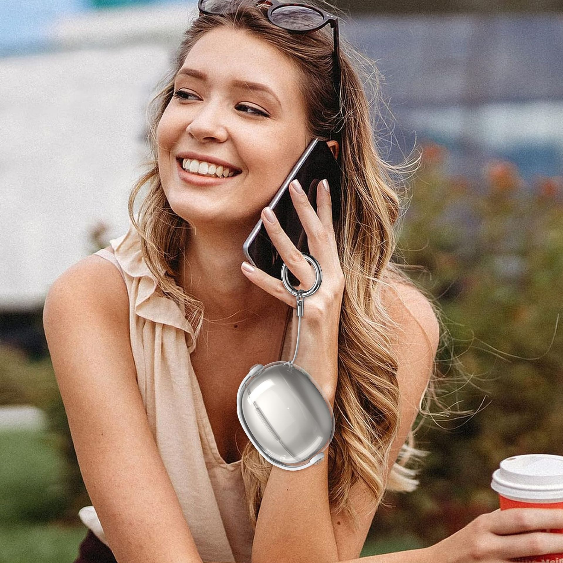 Eine lächelnde Frau mit Brille auf dem Kopf telefoniert mit einem silbernen Ohrhörer-Etui, das an einem Ring an ihrem Finger befestigt ist.