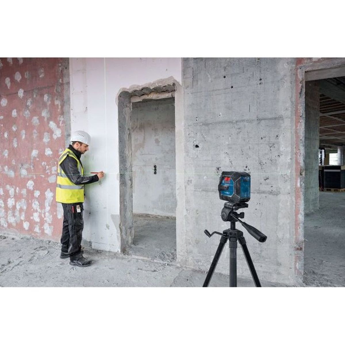 Ein Bauarbeiter in Helm und Weste misst eine Wand mit einem Laser-Wasserwaage auf einem Stativ.