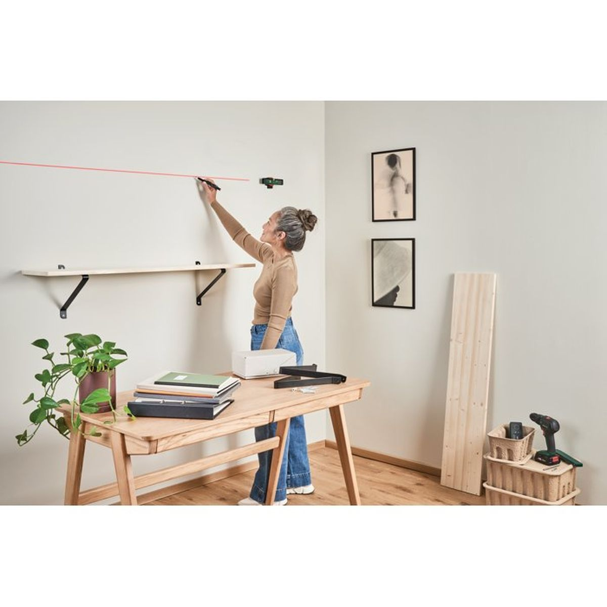 Eine Frau benutzt eine Laserwasserwaage, um eine Linie an einer weißen Wand über einem Holzregal zu markieren. Ein Tisch mit Gegenständen ist im Vordergrund.