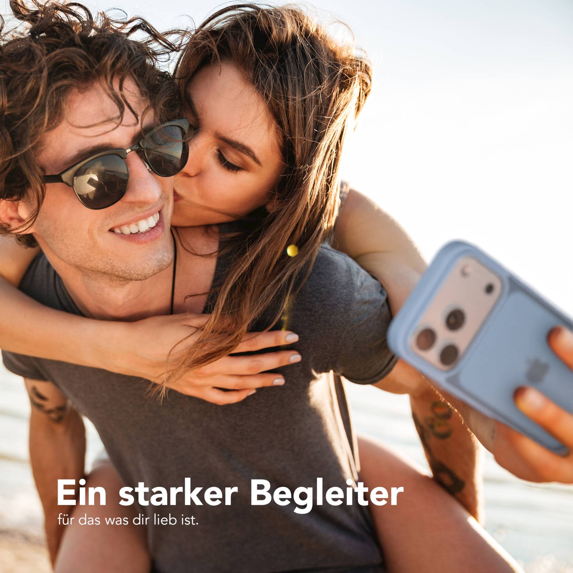 Eine Frau küsst einen Mann auf die Wange, während er lächelt und ein hellblaues Smartphone mit Hülle im Freien hält.
