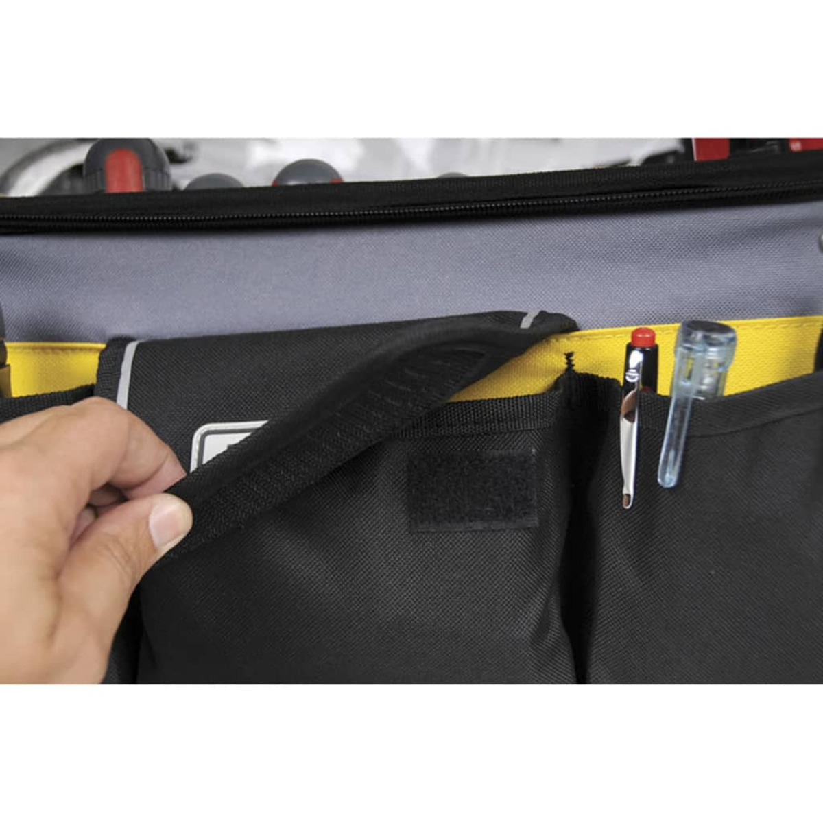 Eine Hand öffnet eine schwarze Werkzeugtasche mit gelbem Innenfutter, Stifte und Werkzeuge sind in Taschen vor grauem Hintergrund zu sehen.