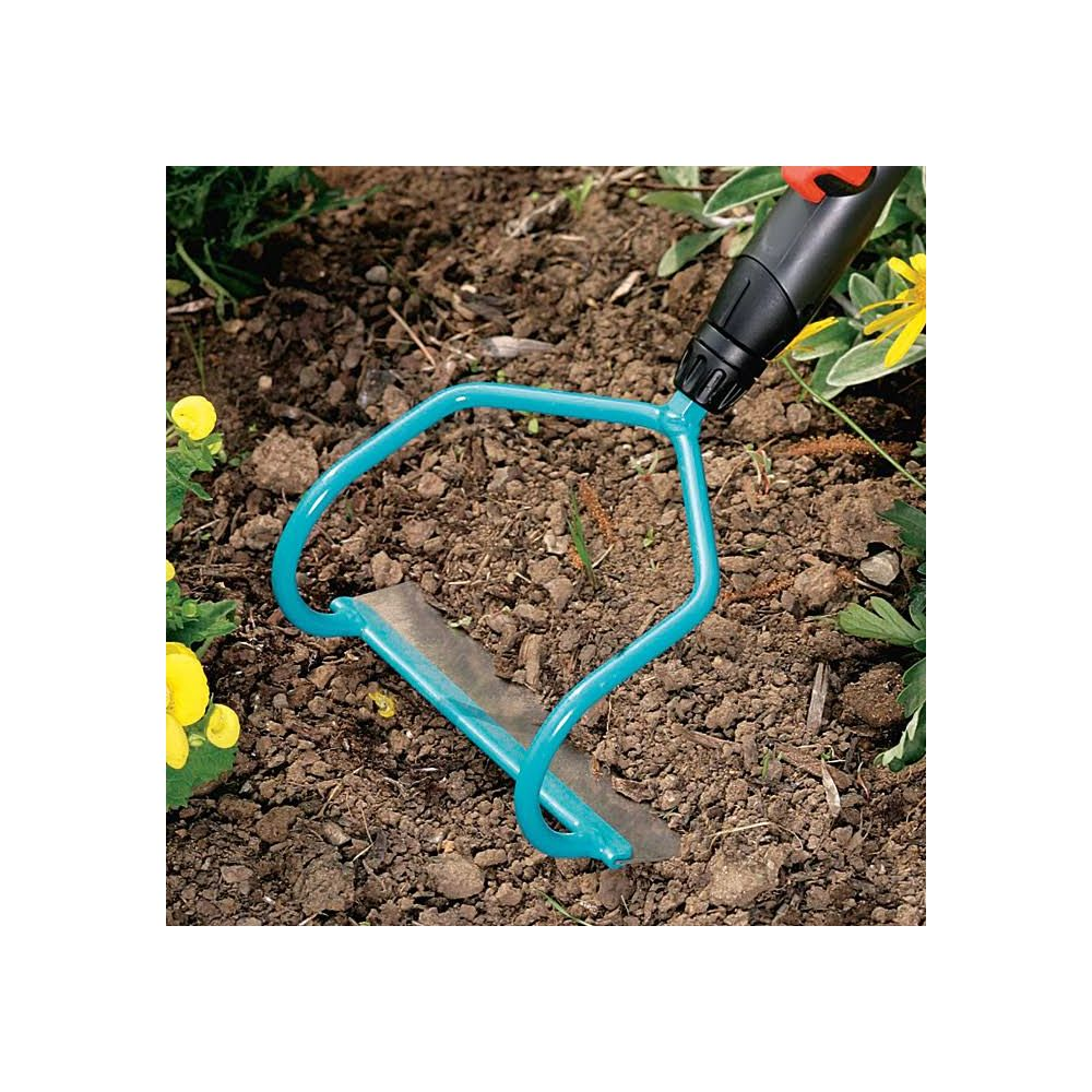 Eine Nahaufnahme zeigt eine türkisfarbene Gartenhacke in dunkelbrauner Erde mit gelben Blumen und grünem Laub im Hintergrund.