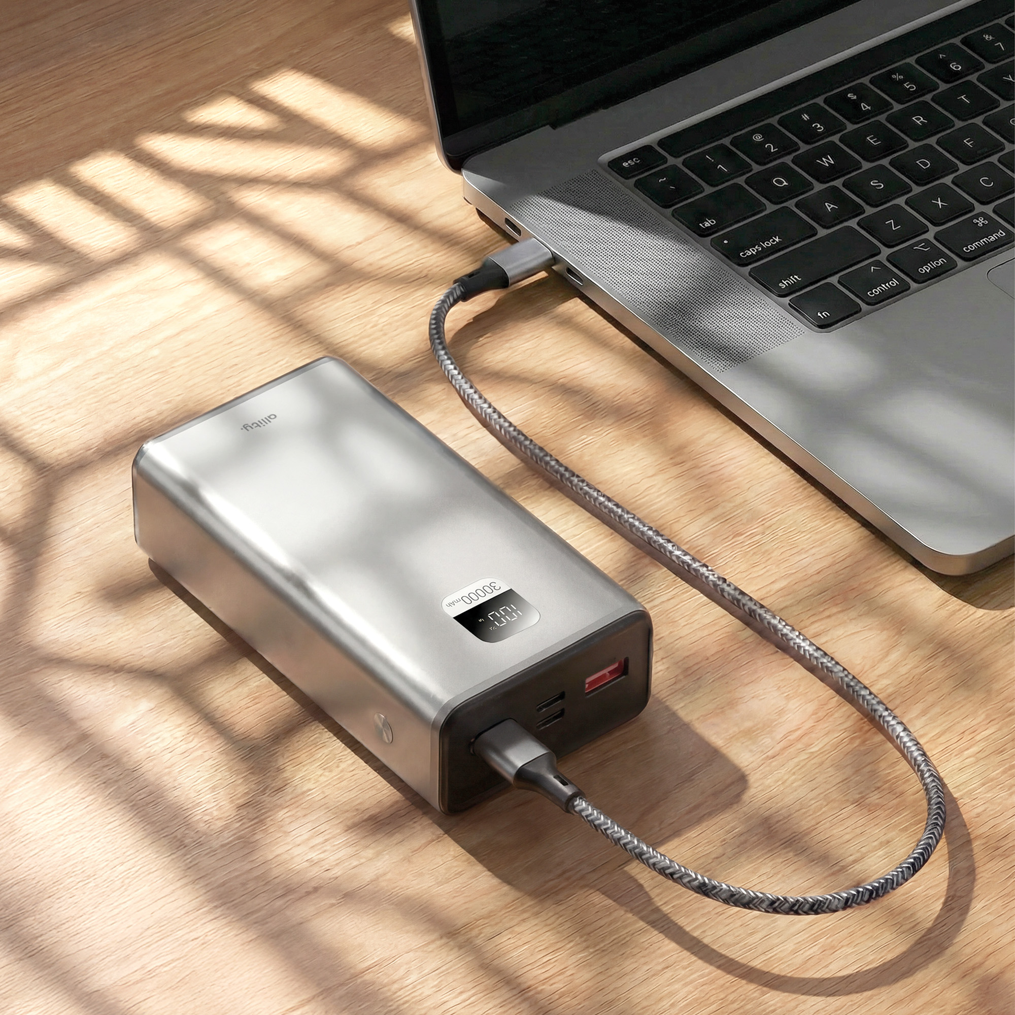 Eine silberne Powerbank, die mit einem geflochtenen Kabel an einen grauen Laptop auf einem Holztisch mit Sonnenstreifen angeschlossen ist.