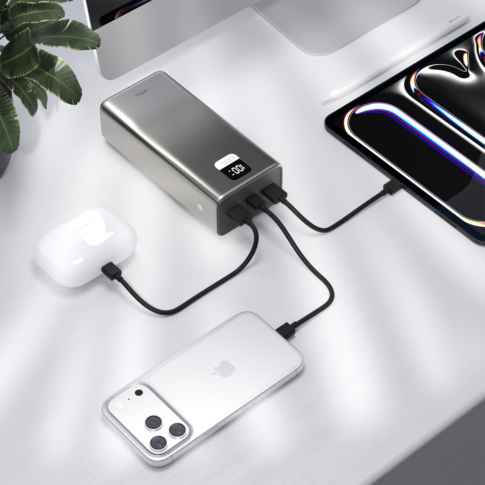 Eine graue Powerbank lädt ein weißes iPhone, AirPods und ein Tablet auf einem weißen Schreibtisch mit einer Pflanze.
