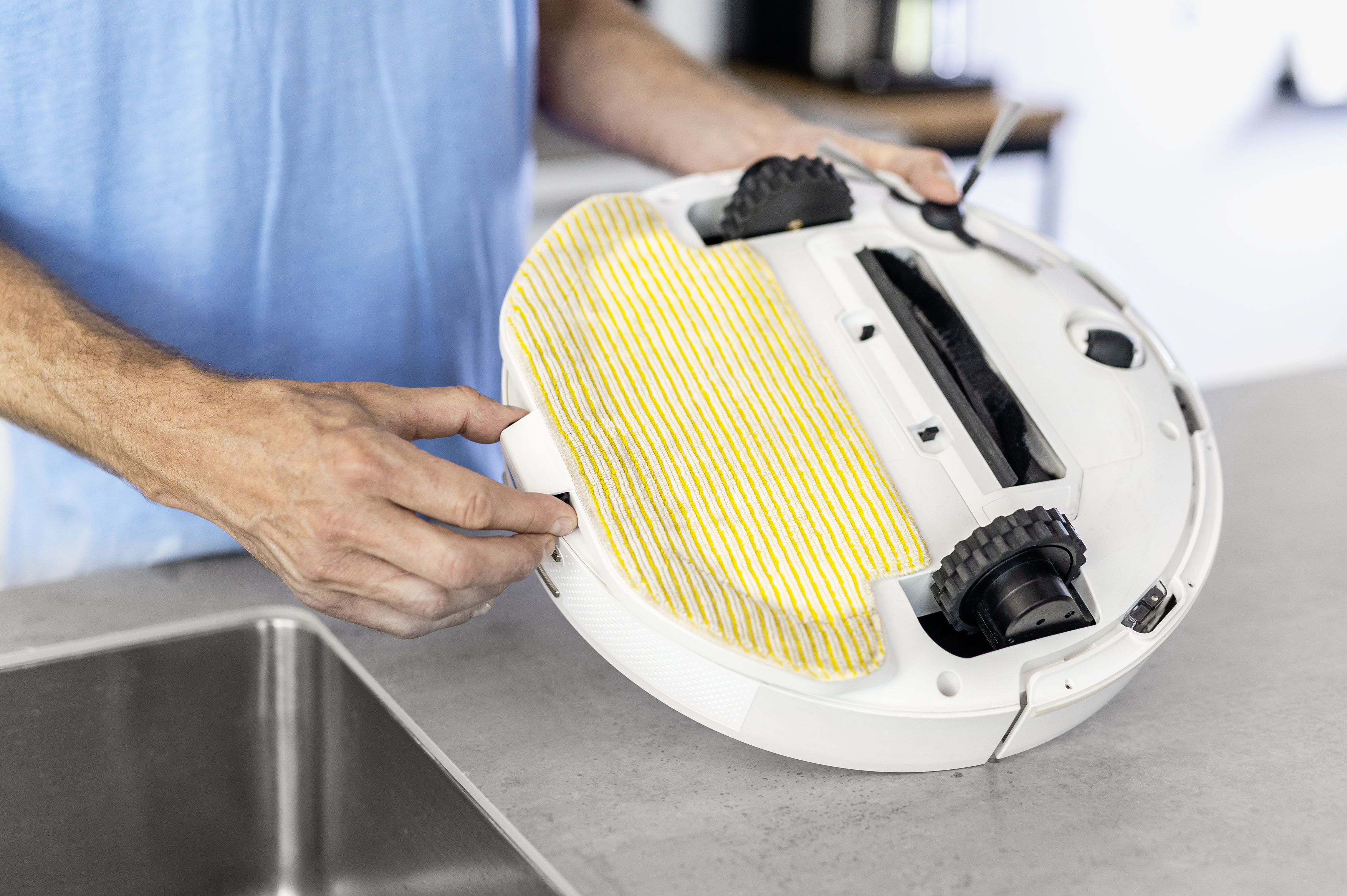 Una persona con una maglietta blu tiene un robot aspirapolvere bianco capovolto, rivelando un panno mop a strisce gialle e bianche.