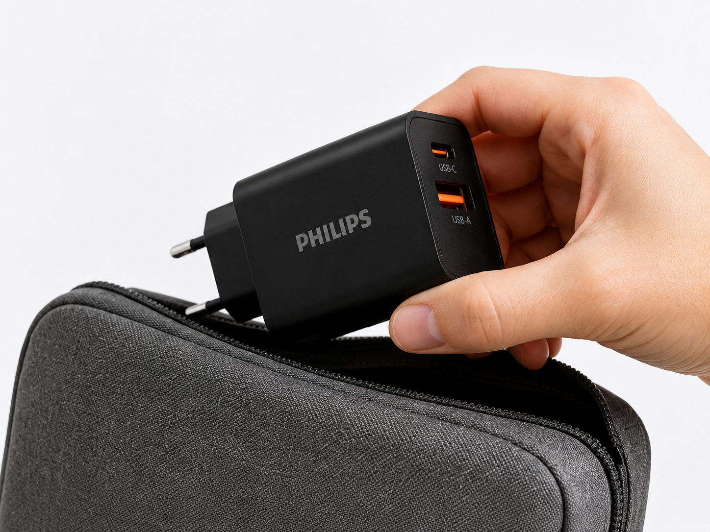 Bir el, USB-C ve USB-A bağlantı noktalarına sahip siyah bir Philips şarj cihazını beyaz bir arka plan üzerinde gri fermuarlı bir keseye yerleştiriyor.