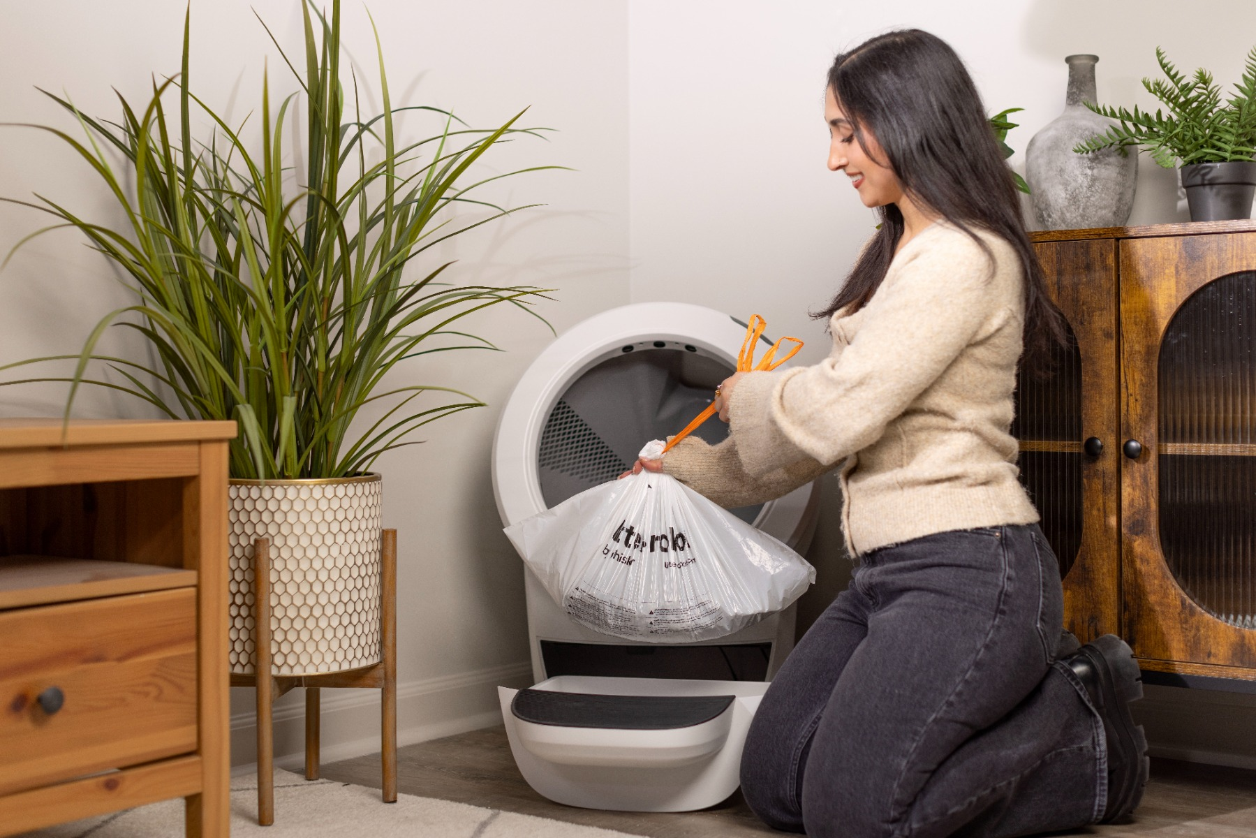 Mujer arrodillada, sosteniendo una bolsa de basura llena con pinzas naranjas, junto a una caja de arena automática blanca.