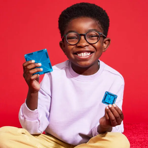 Un garçon souriant portant des lunettes tient des blocs de construction magnétiques bleus sur fond rouge.