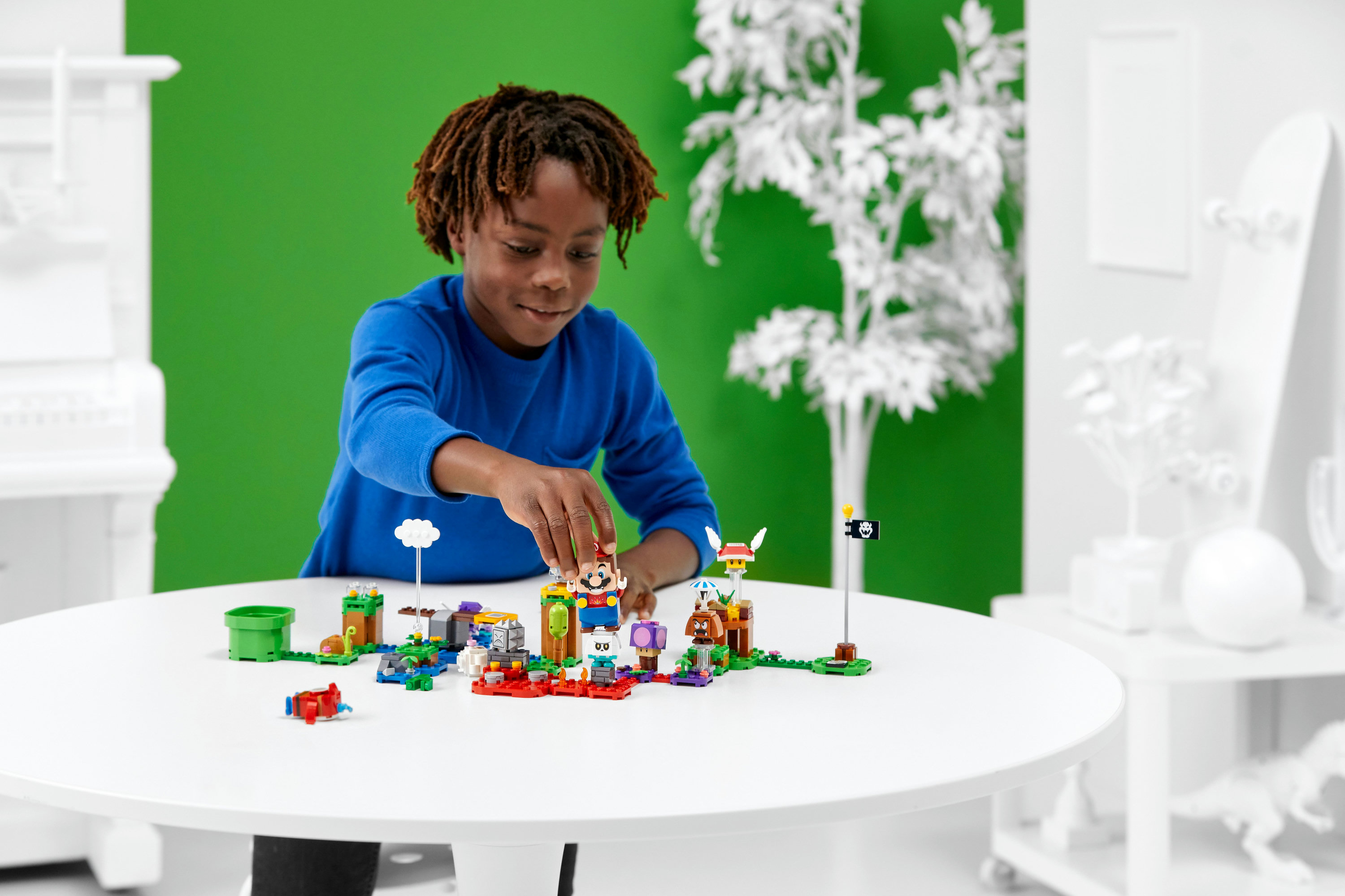 Un garçon aux dreadlocks en chemise bleue joue avec un set LEGO Mario coloré sur une table blanche devant un fond vert.