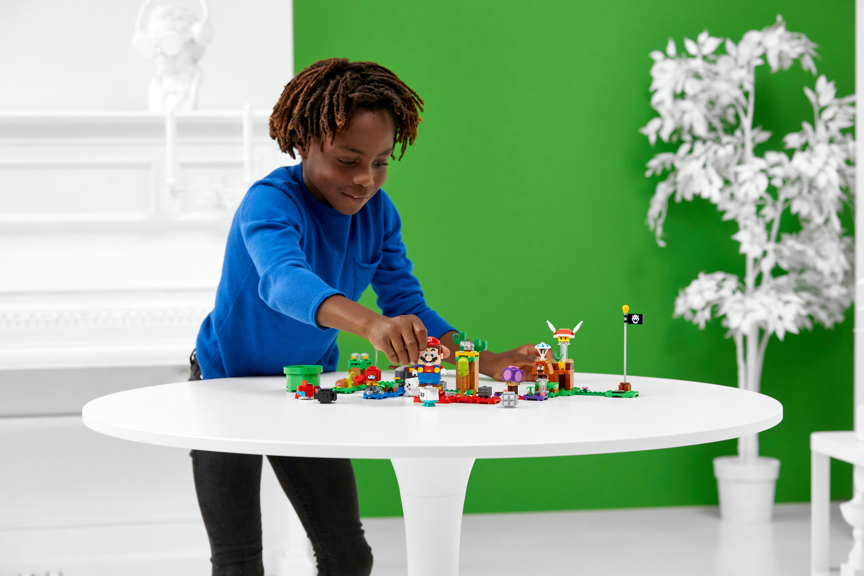 Un garçon aux dreadlocks en chemise bleue joue avec un set Lego Mario coloré sur une table blanche devant un fond vert.