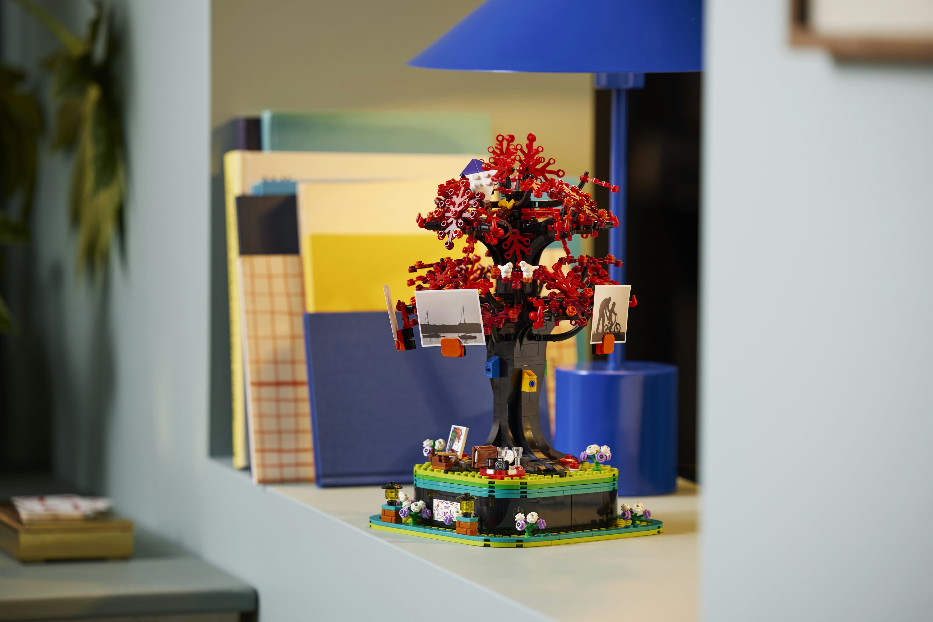 Een Lego Bonsai boom met rode bladeren, versierd met kleine foto's, op een witte plank met boeken en een blauwe lamp.
