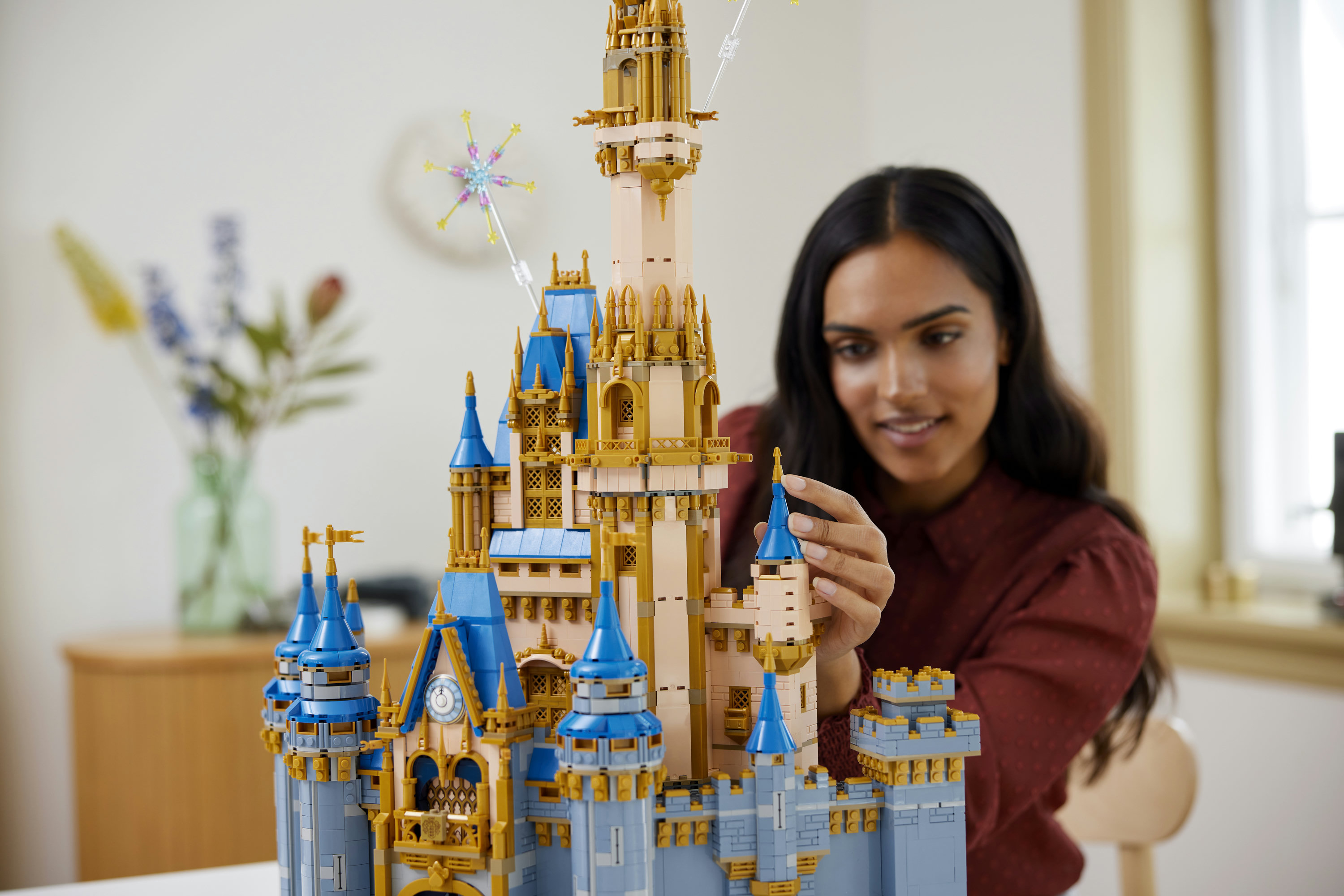 Una donna costruisce un castello Lego beige e blu con accenti dorati. Sullo sfondo un vaso di fiori.