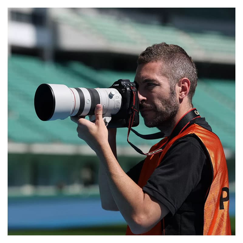Un uomo con la barba scatta una foto con una grande macchina fotografica. Indossa un gilet arancione.