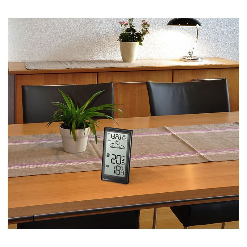 Estación meteorológica digital sobre una mesa de madera con plantas y sillas.