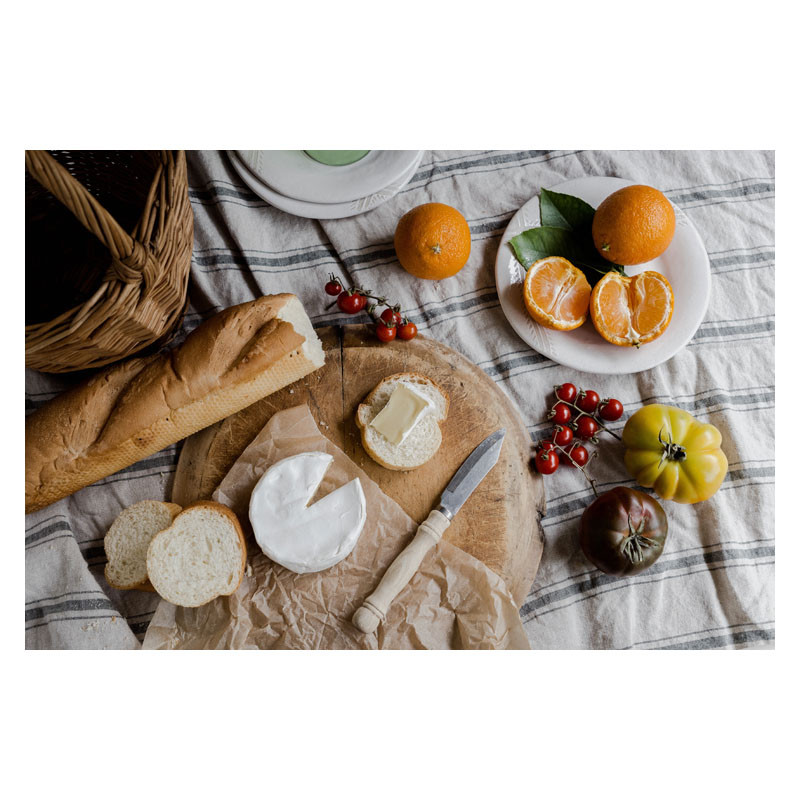 Una baguette, queso, naranjas y tomates están sobre una tela a rayas.