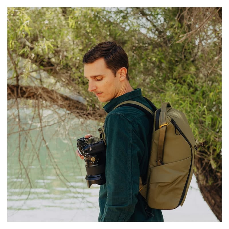 Man with camera and backpack by a river, wearing a dark green shirt.