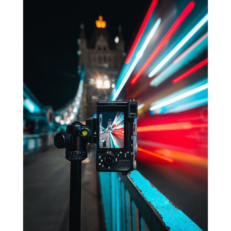 Una cámara en un trípode filma un puente de noche con estelas de luz.