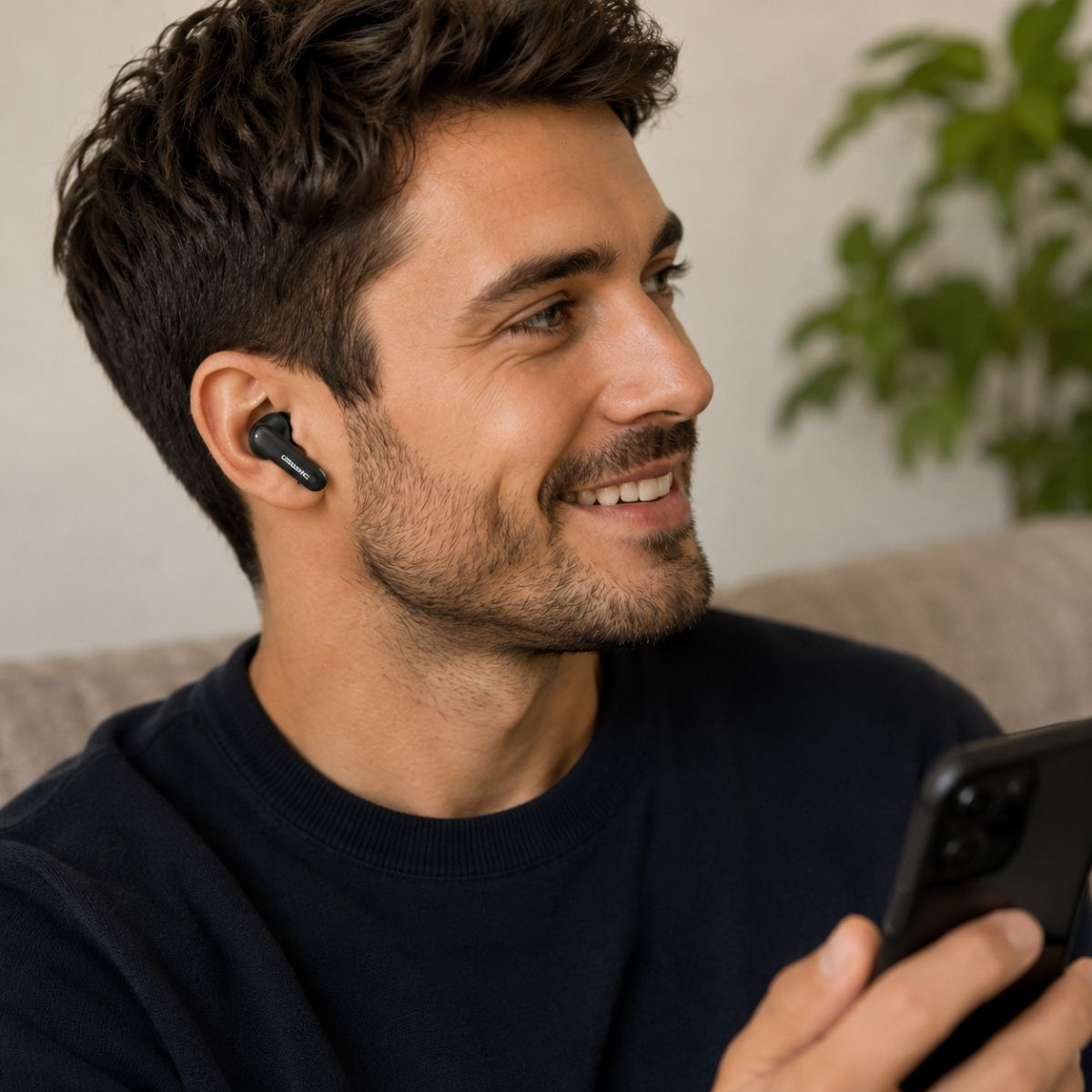 Mann mit schwarzen Ohrhörern lächelt und hält ein Handy. Verschwommener Hintergrund mit Couch und Pflanze.